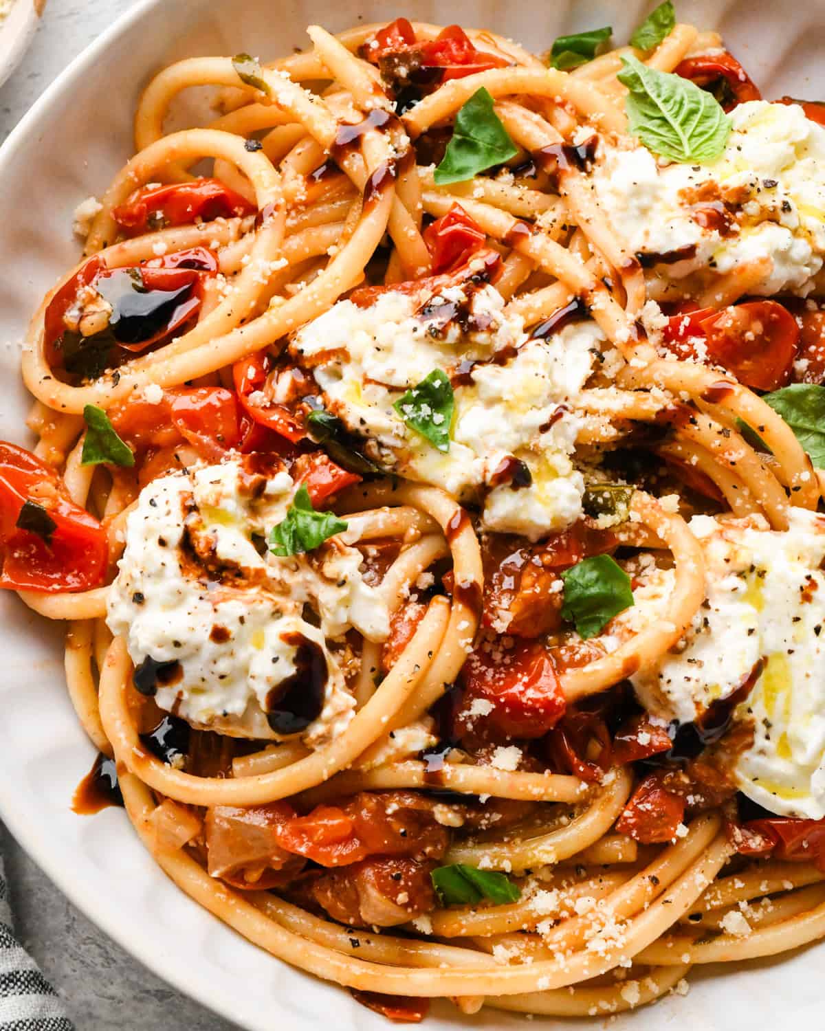 Close-up photo of a white bowl of pasta tossed with roasted tomatoes and fresh basil, topped with creamy burrata cheese and a drizzle of balsamic glaze.
