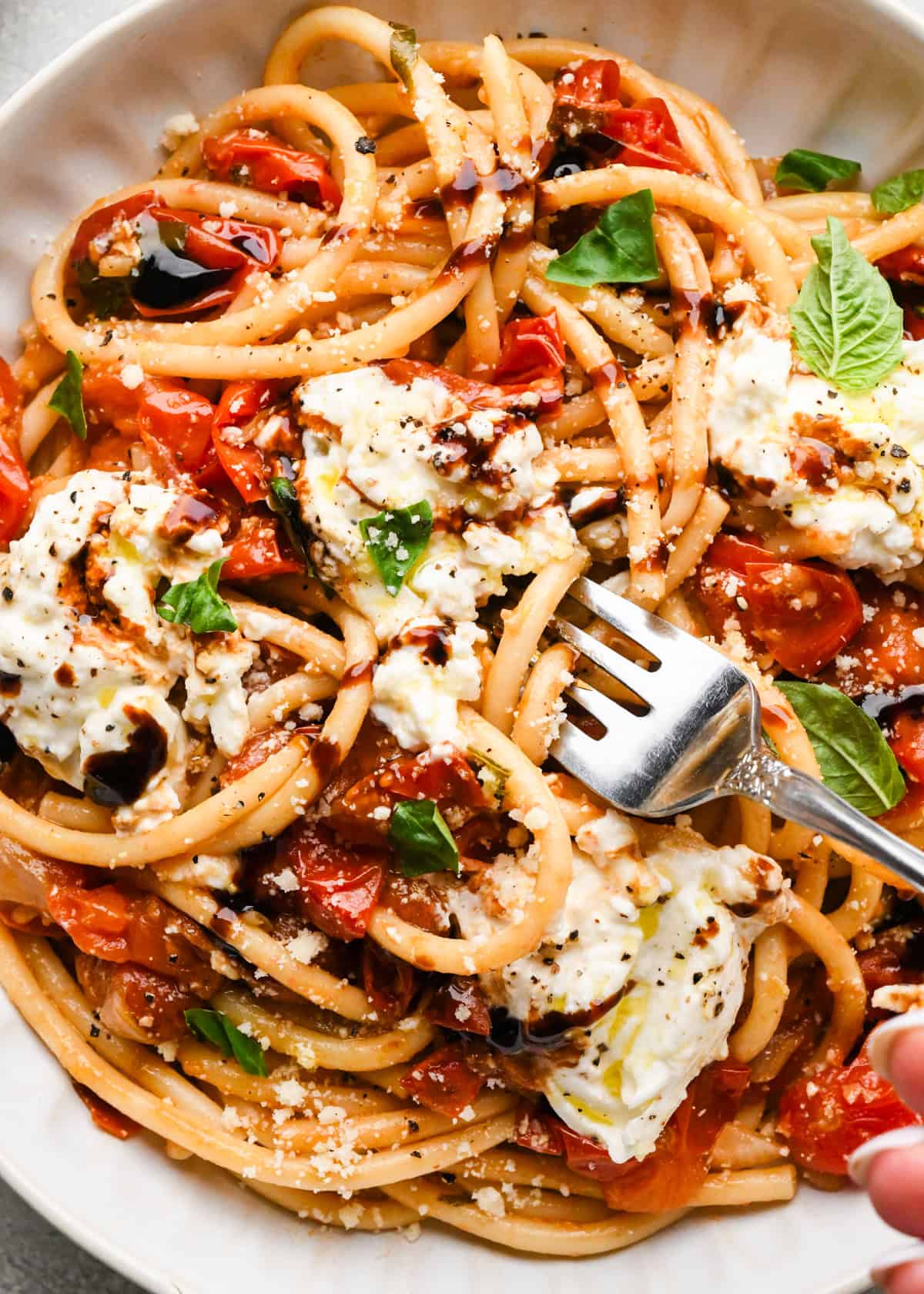 Close-up photo of a white bowl of pasta tossed with roasted tomatoes and fresh basil, topped with creamy burrata cheese and a drizzle of balsamic glaze with a fork resting in the bowl.