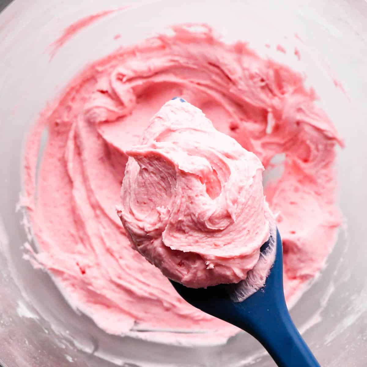 a blue spatula holding a scoop of raspberry frosting over a glass bowl of raspberry frosting