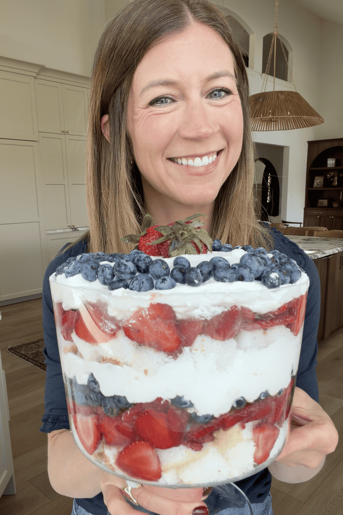laura from joyfoodsunshine smiling and holding a fresh berry trifle in a trifle dish with blueberries, strawberries, whipped cream and angel food cake