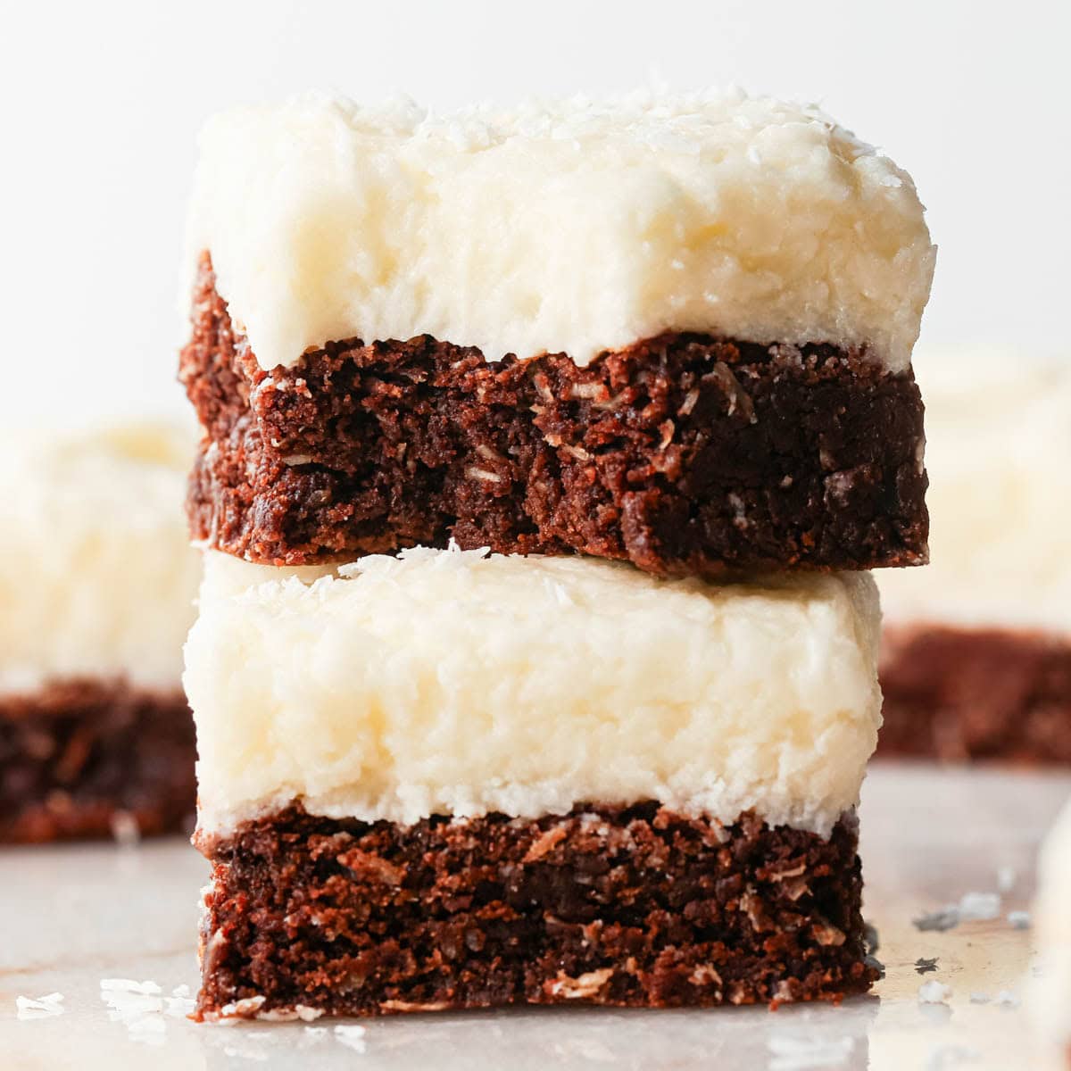 a stack of two chocolate coconut brownies with coconut frosting, the top one has a bite taken out of it and there are more brownies in the background