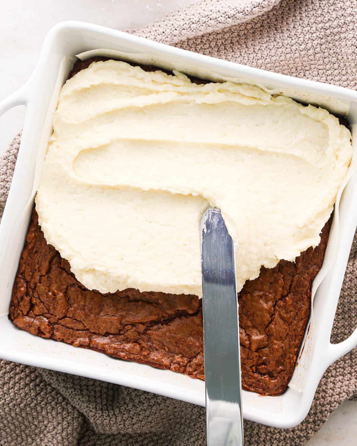 coconut frosting being spread over coconut brownies in a white baking dish