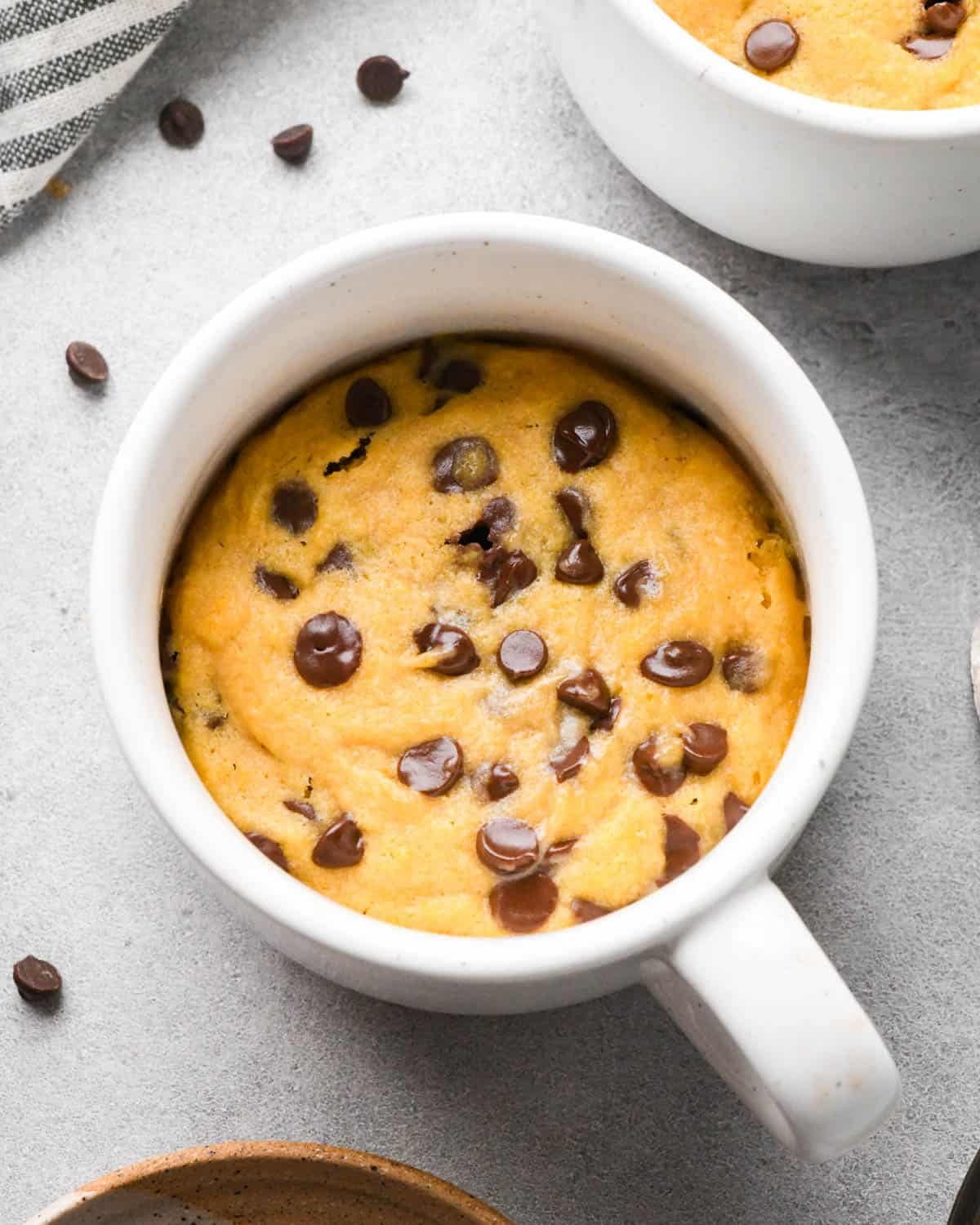 chocolate chip mug cookie in a white mug with more chocolate chips, a kitchen towel and another mug cookie in the background. 