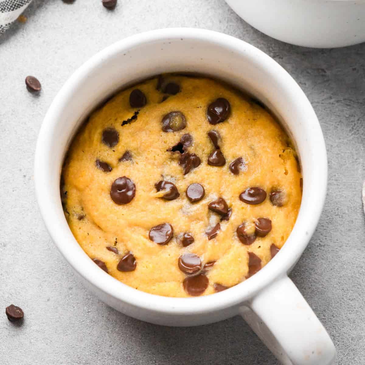 chocolate chip mug cookie in a white mug with more chocolate chips, a kitchen towel and another mug in the background. 
