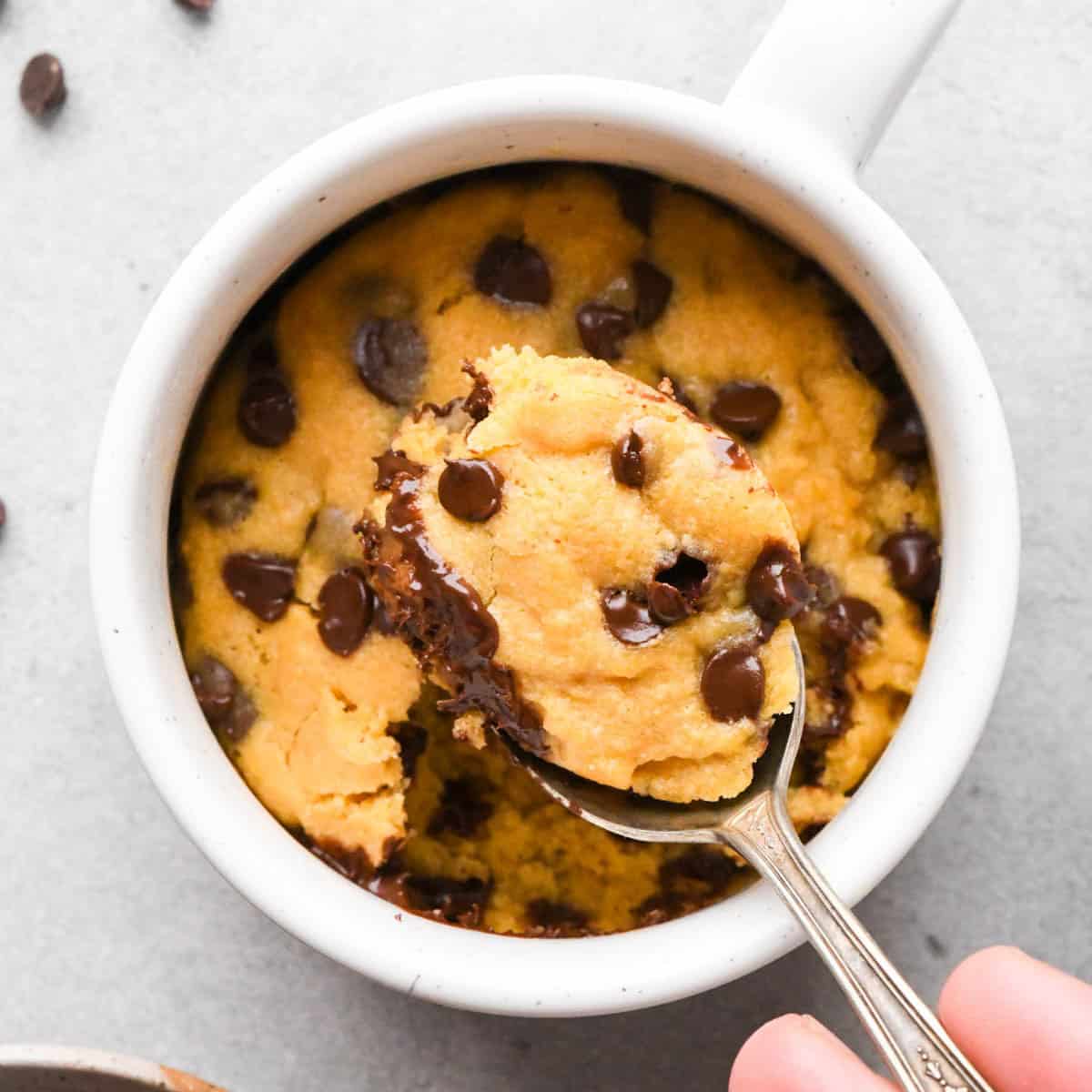a spoon holding a scoop of mug cookie over a chocolate chip cookie in a mug 