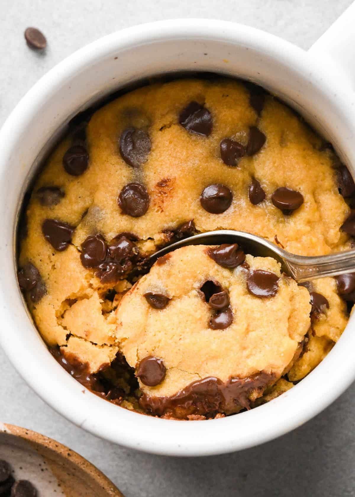 a silver spoon taking a scoop out of a chocolate chip mug cookie in a white mug with more chocolate chips, a kitchen towel and another mug in the background. 