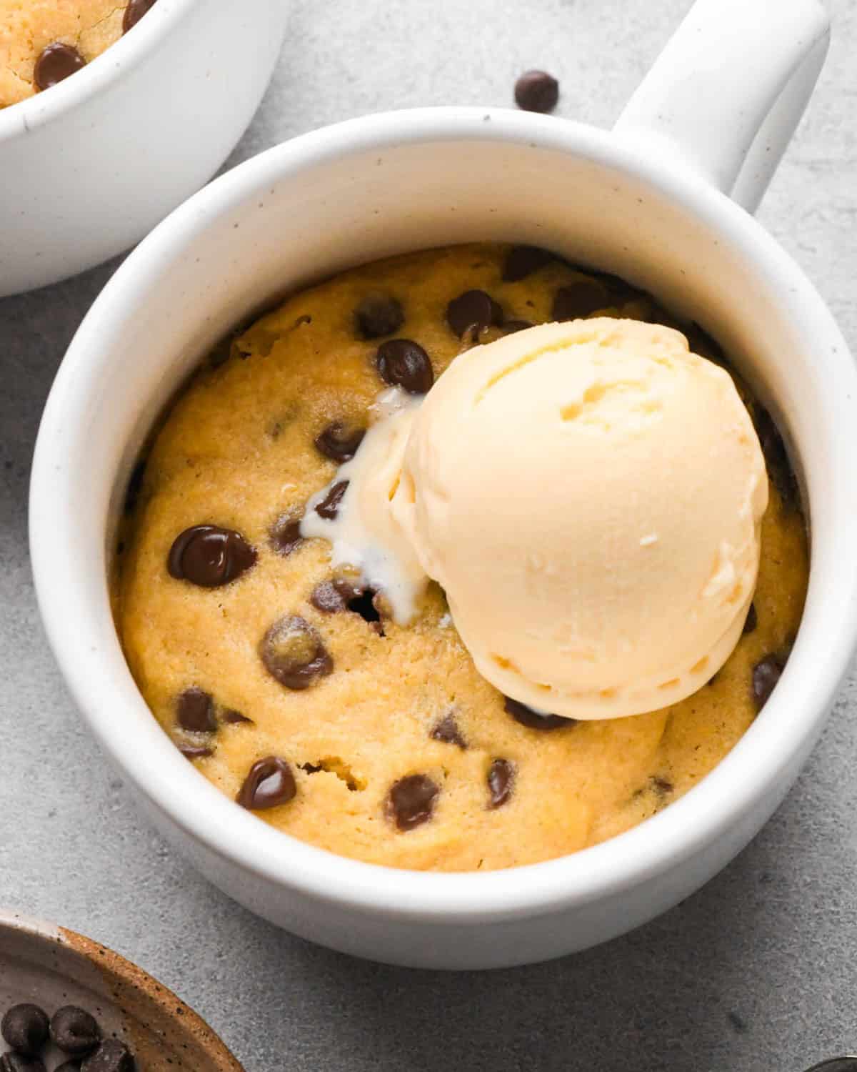 chocolate chip mug cookie in a white mug topped with some vanilla ice cream, with more chocolate chips and another mug cookie in the background. 