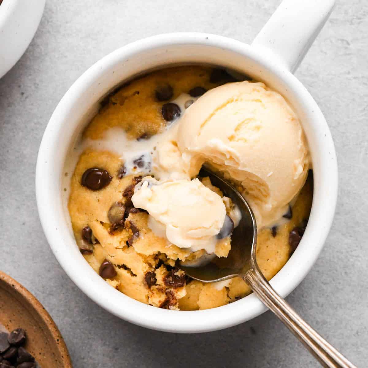 a spoon taking a scoop out of a chocolate chip mug cookie in a white mug topped with some vanilla ice cream, with more chocolate chips and another mug in the background. 