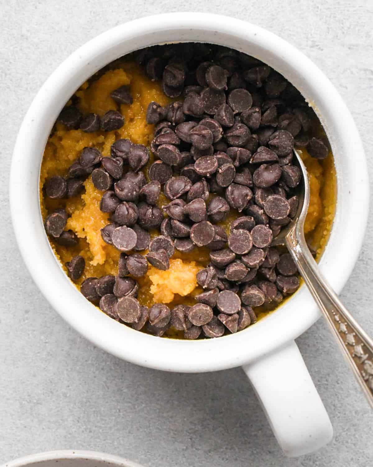 making a cookie in a mug - chocolate chips added to a white mug with a silver spoon before mixing