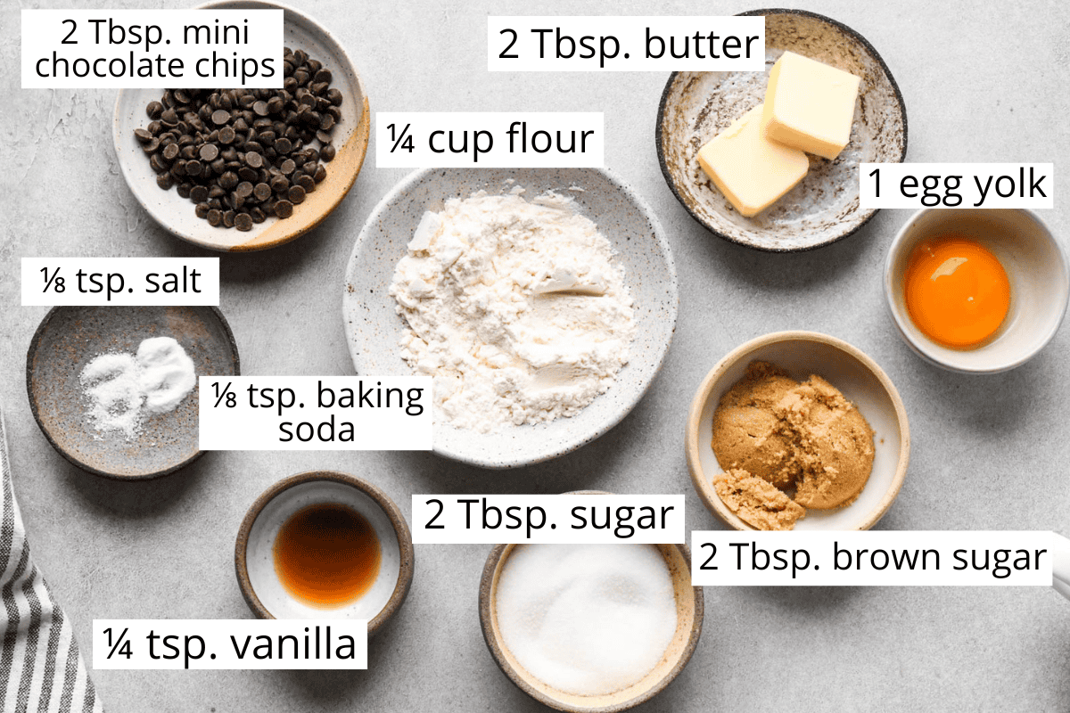 overhead photo of the ingredients for this mug cookie recipe in bowls and labeled with what they are and the amounts like butter, flour, egg yolk, sugar, chocolate chips, etc. 