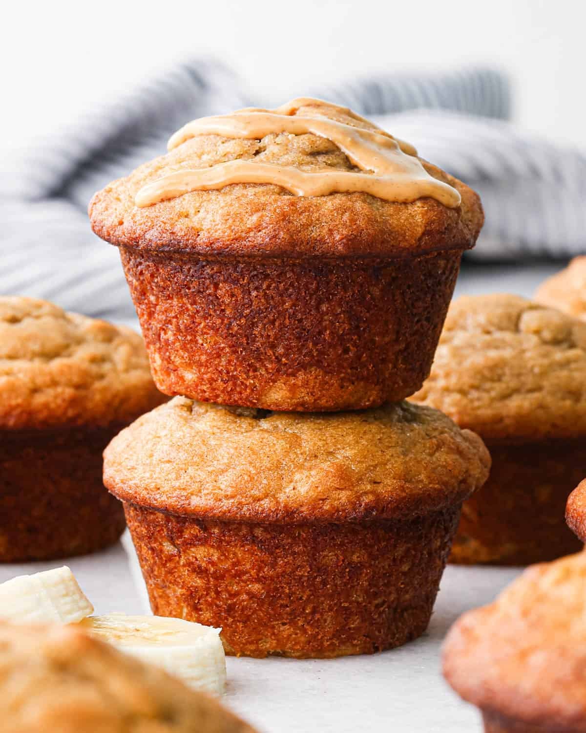 a stack of two peanut butter banana muffins, the top one has peanut butter drizzled on top. There are other muffins, sliced bananas and peanut butter surrounding the muffin stack.