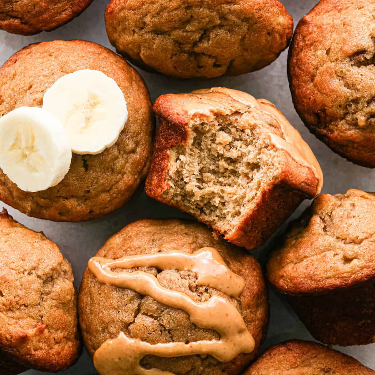 7 peanut butter banana muffins, one laying on it's side with a bite taken out so the moist texture is showing, it also has peanut butter drizzled on top