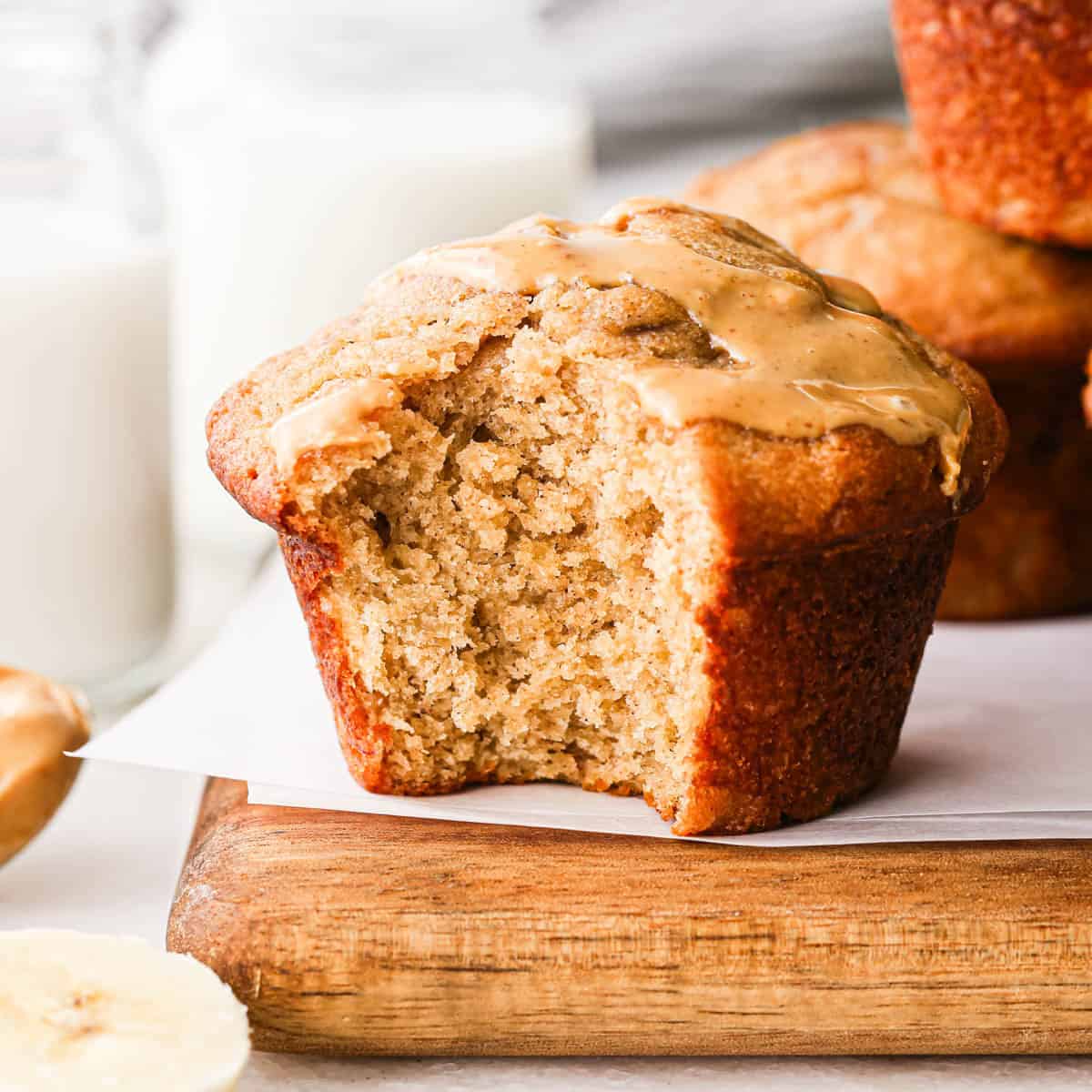 a peanut butter banana muffin with a bite taken out of it and peanut butter drizzled on top. There is a spoon of peanut butter and a stack of two muffins in the background