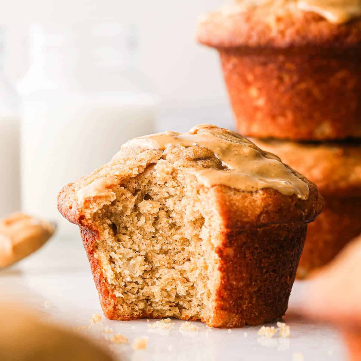a peanut butter banana muffin with a bite taken out of it and peanut butter drizzled on top. There is a spoon of peanut butter and a stack of two muffins in the background