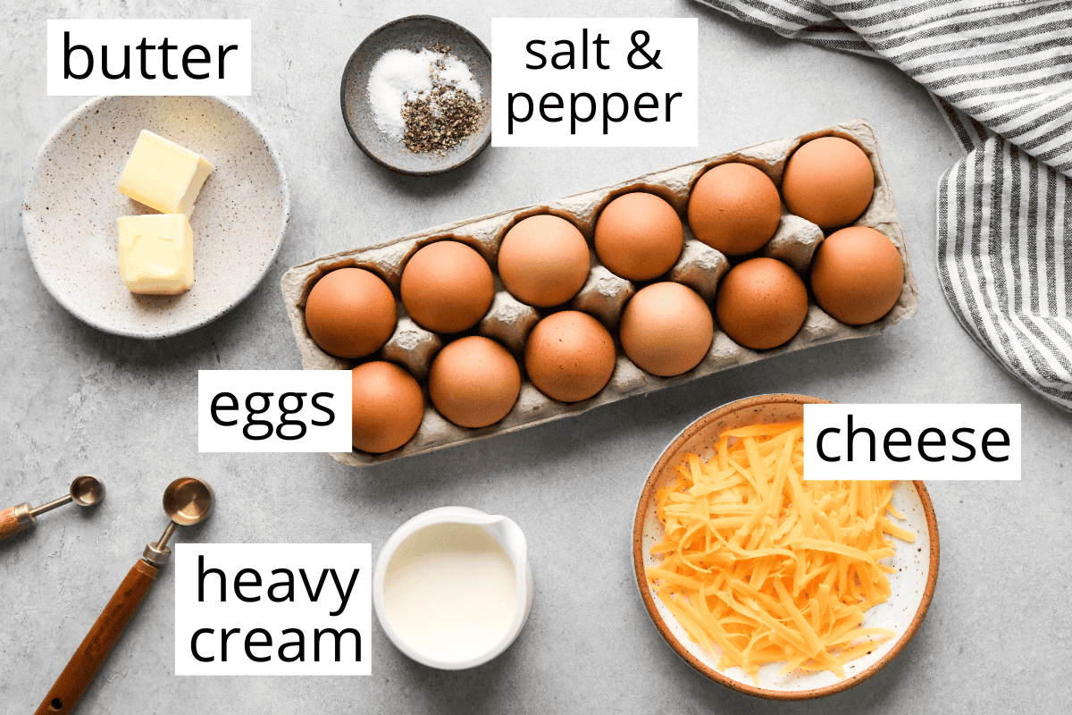 overhead photo of the ingredients in this baked scrambled eggs recipe all labeled, 12 eggs in a carton, then the rest are in bowls including cheese, cream, butter salt and pepper