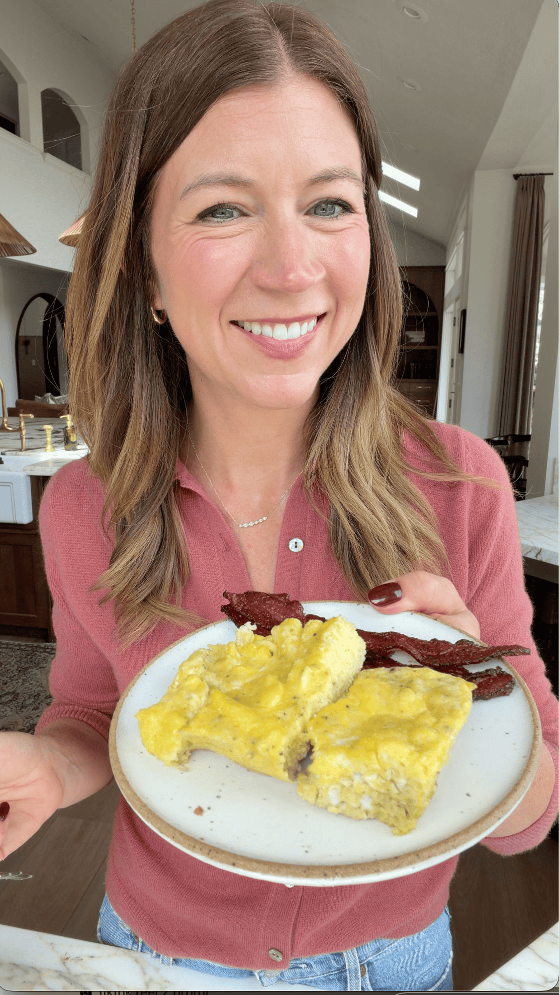 a photo of laura from joyfoodsunshine in a mauve pink sweater smiling holding a white ceramic plate with baked scrambled eggs recipe on it along with a side of bacon