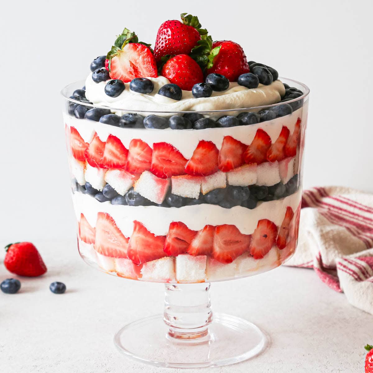 a berry trifle in a glass trifle dish with 3 layers of each: angel food cake, strawberries, whipped cream and blueberries. There is a red and cream striped towel and berries in the background. It's topped with strawberries, blueberries and whipping cream