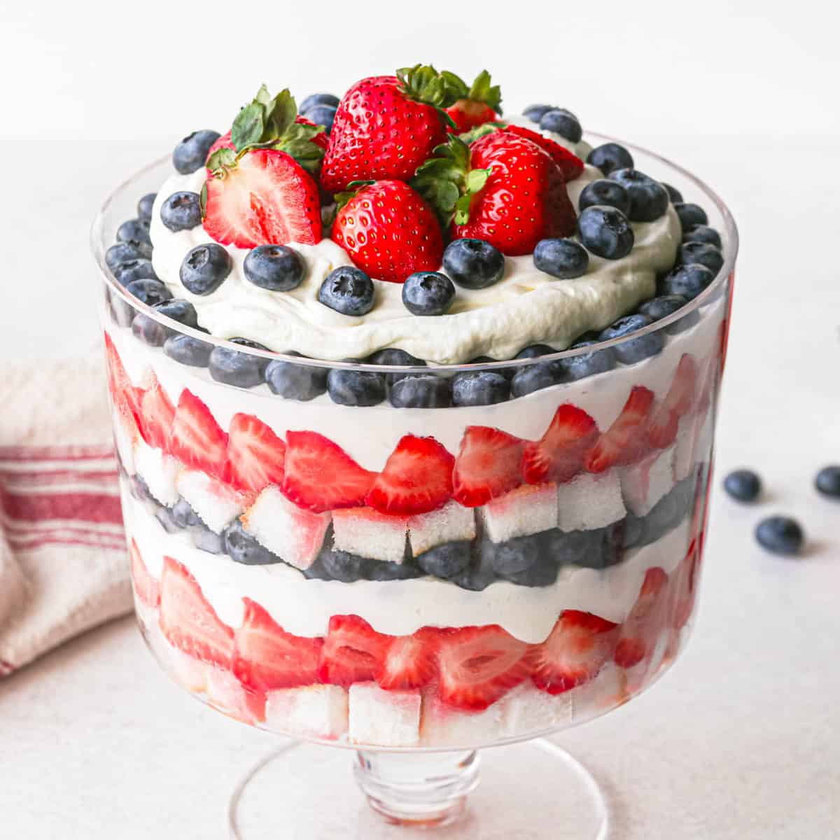 a berry trifle in a glass trifle dish with 3 layers of each: angel food cake, strawberries, whipped cream and blueberries. There is a red and cream striped towel and blueberries in the background. It's topped with strawberries, blueberries and whipping cream