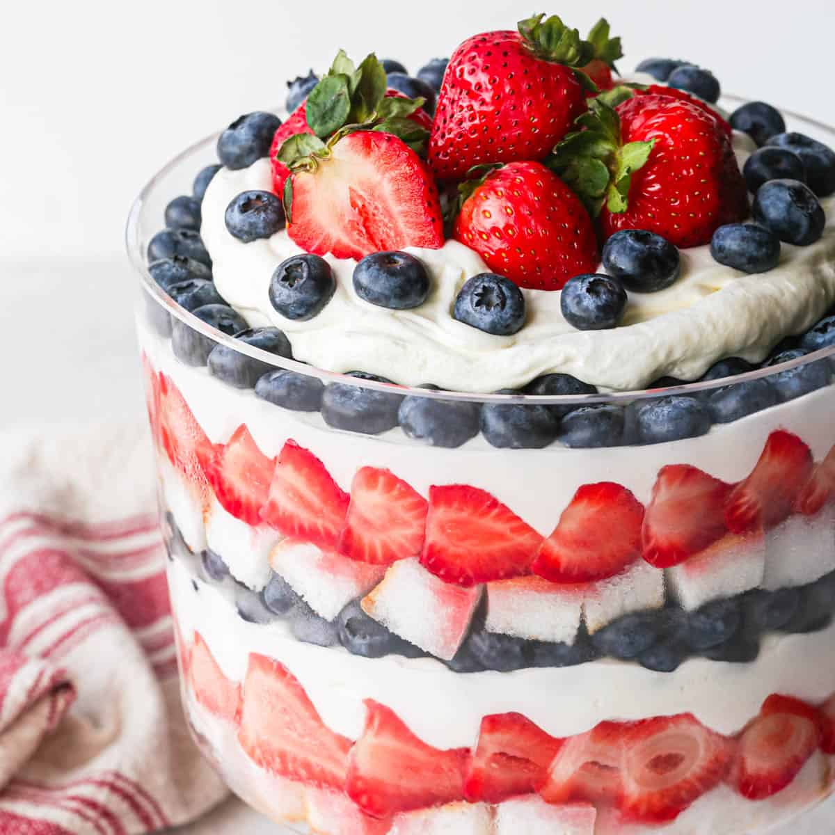 up close photo of a berry trifle in a glass trifle dish with 3 layers of each: angel food cake, strawberries, whipped cream and blueberries. There is a red and cream striped towel in the background around the trifle dish