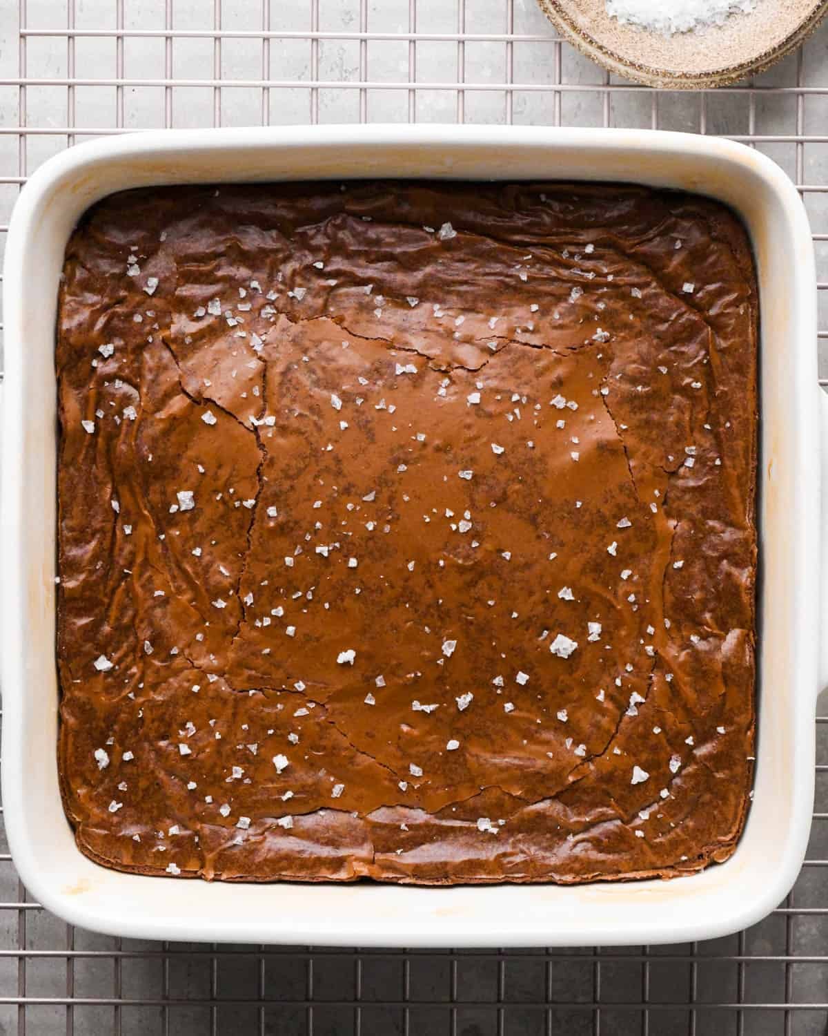 baked brown butter brownies in a white square baking dish on a wire cooling rack topped with flaky sea salt