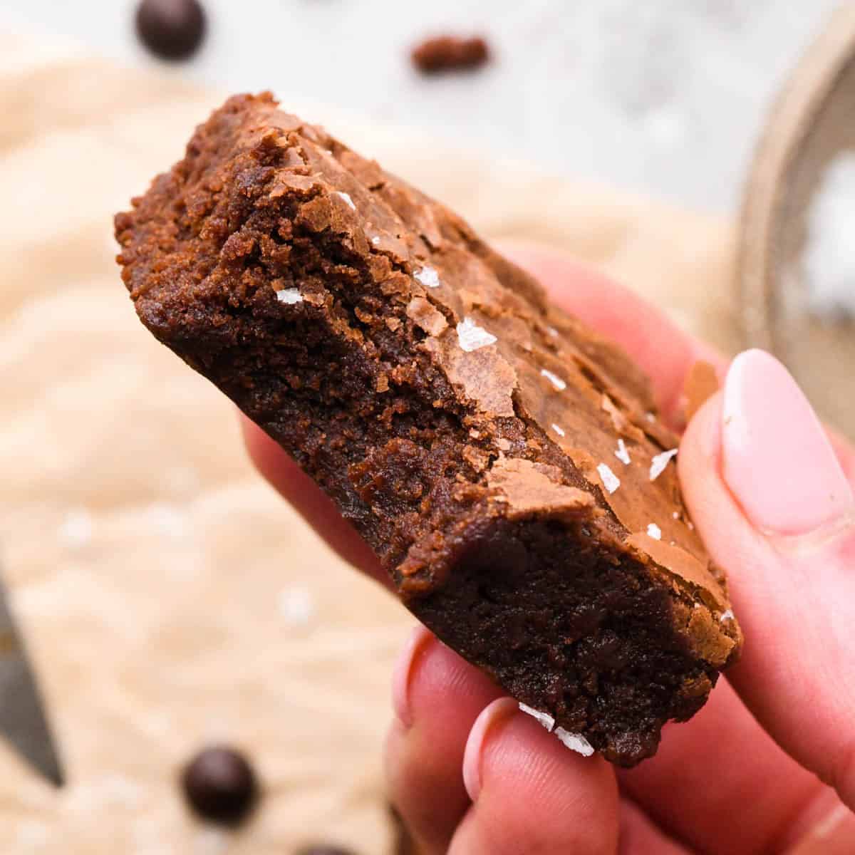 a hand holding a browned butter brownie topped with flaky sea salt with a bite taken out of it to show the fudgy texture inside