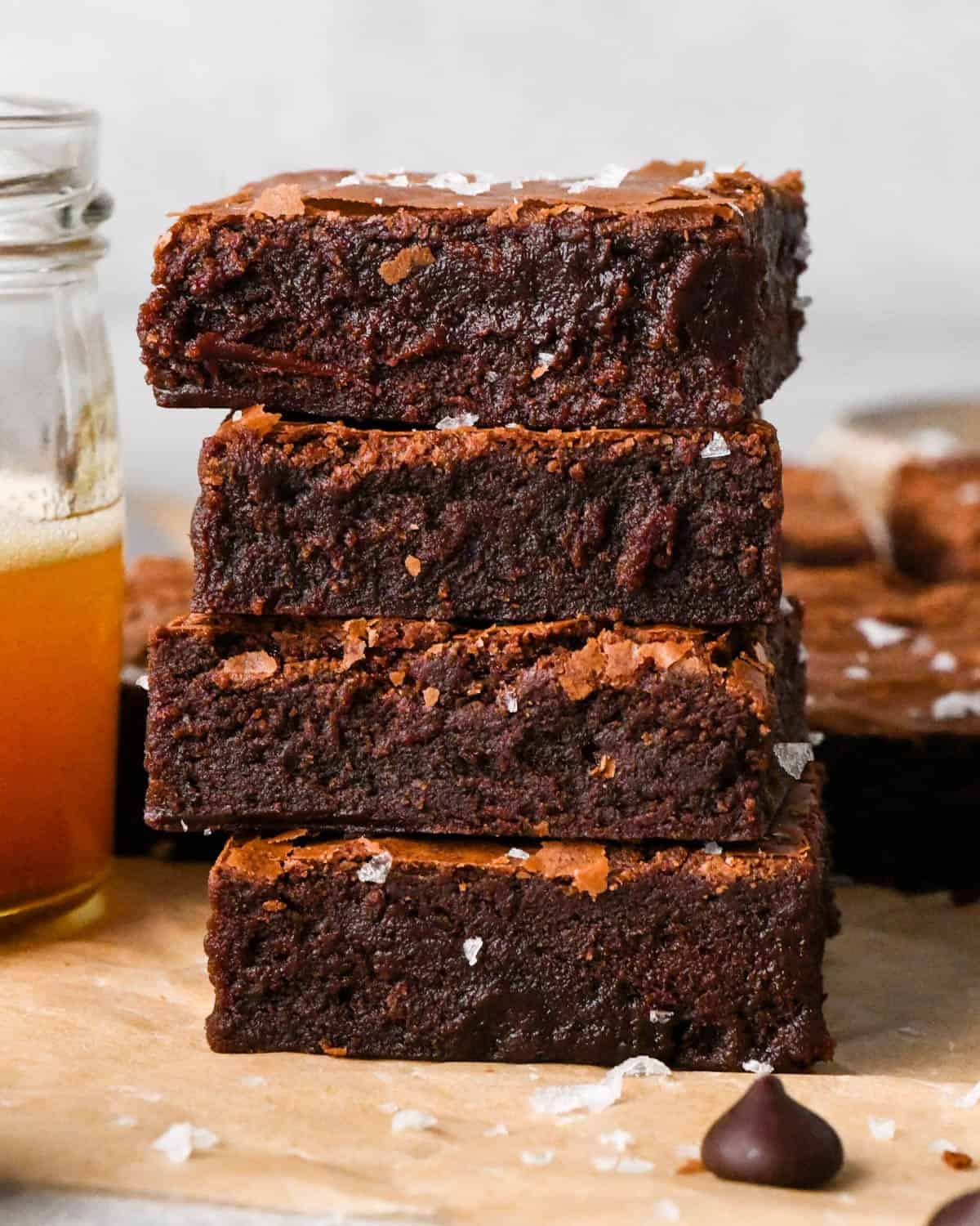 a stack of four fudgy brown butter brownies with flaky sea salt on parchment paper with more brownies and a jar of browned butter in the background