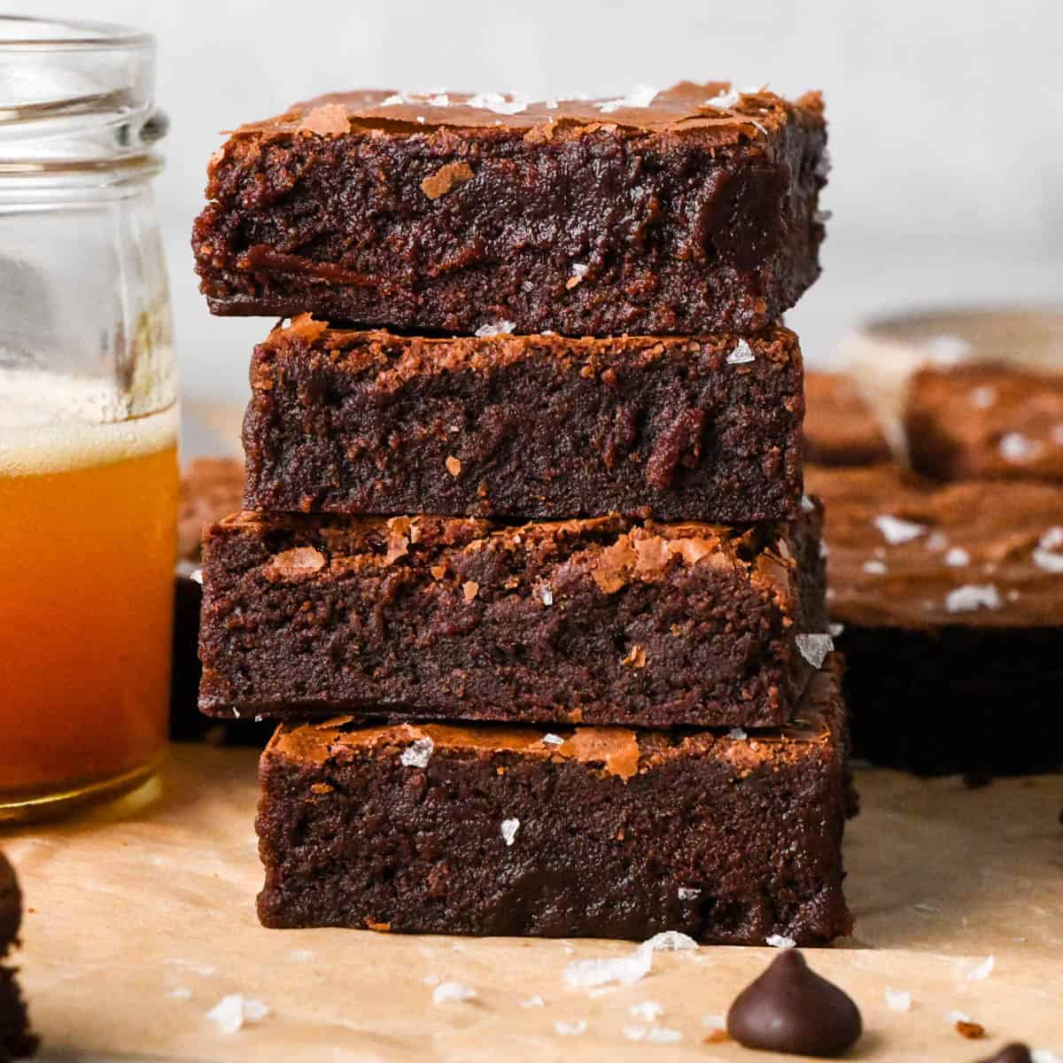 a stack of 4 brown butter brownies with flaky sea salt on top and their fudgy texture showing, there is a jar of brown butter and more brownies in the background.