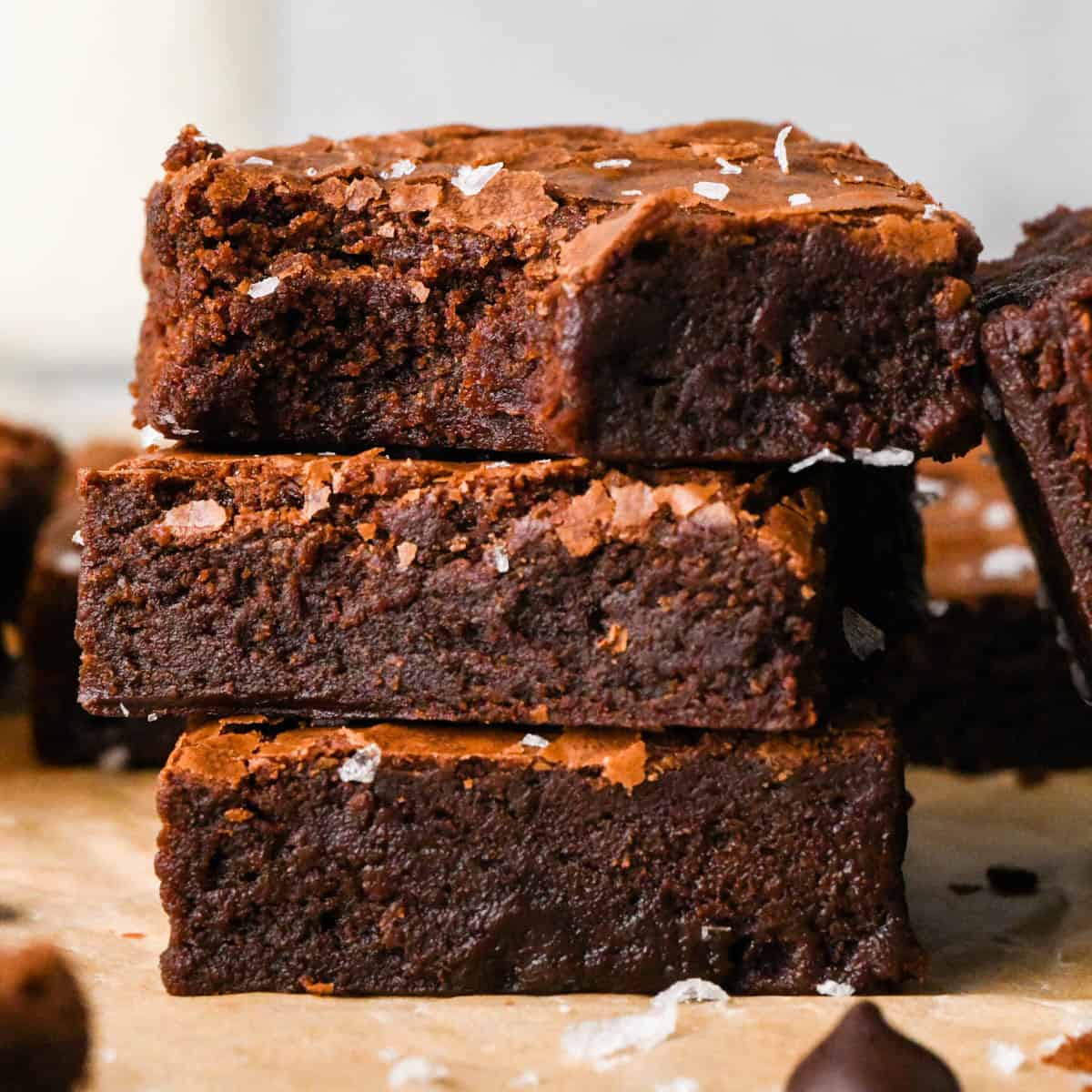a stack of 3 brown butter brownies with flaky sea salt on top and their fudgy texture showing, the top brownie has a bite taken out of it
