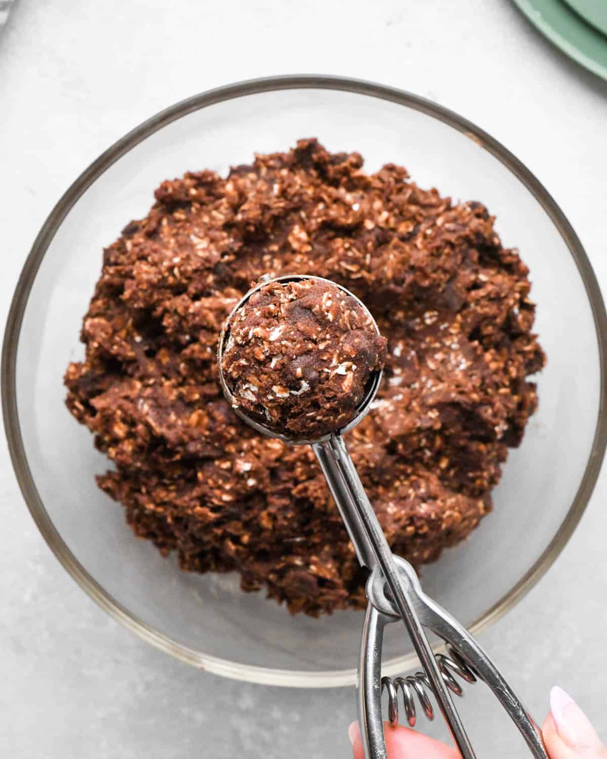 a hand holding a cookie scoop with a portion of final double chocolate oatmeal cookies dough being held over a glass bowl with more dough