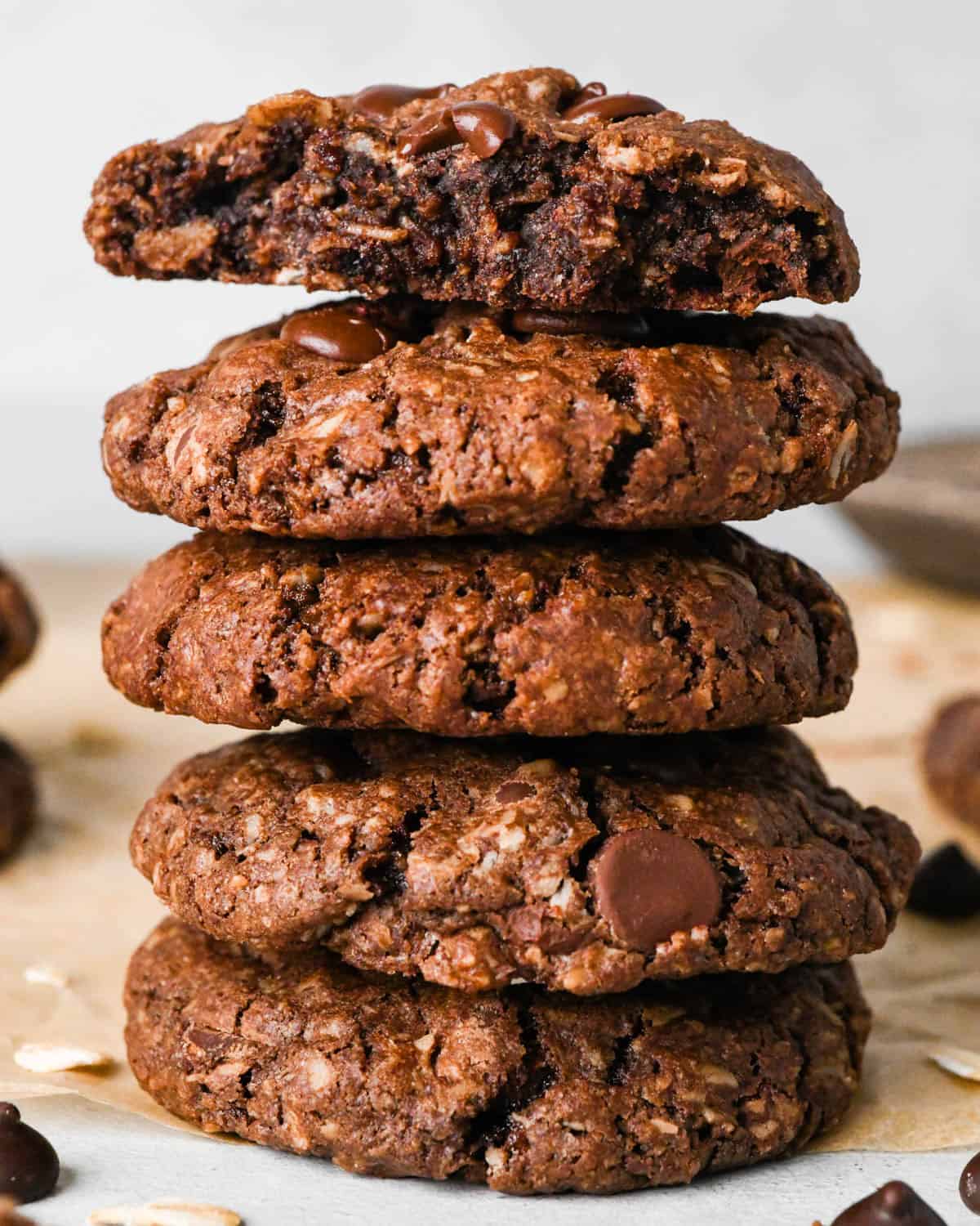 a stack of 5 double chocolate oatmeal cookies on parchment paper surrounded by chocolate chips and oats. The top cookie is cut in half so the gooey chocolatey center is visible