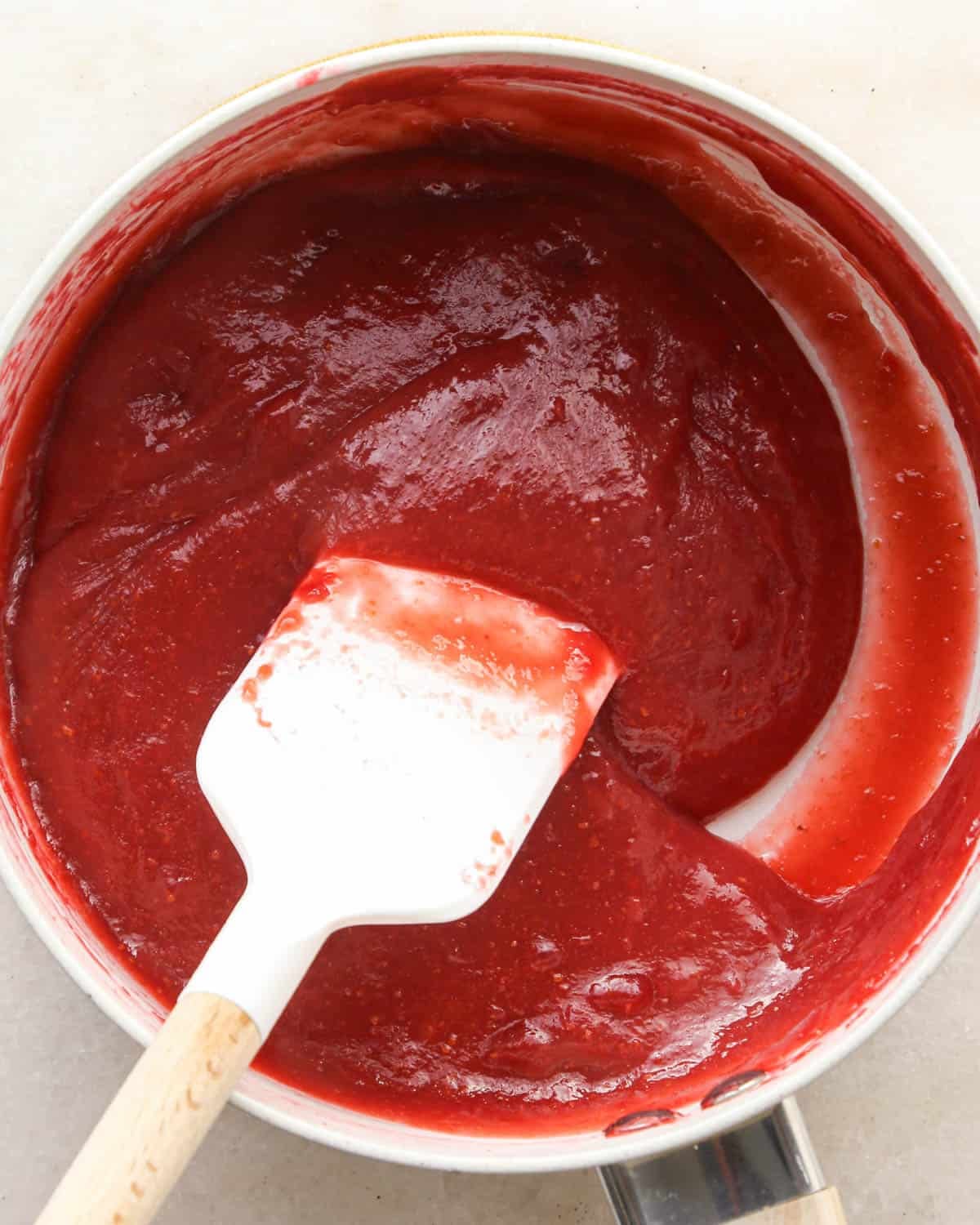 thickened raspberry filling in a white saucepan being stirred with a white spatula. It's thickened texture and bright pink/red color are both visible in this photo