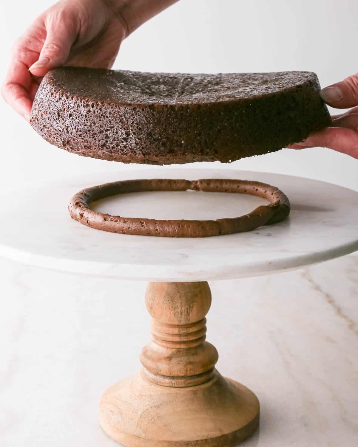 assembling chocolate raspberry cake - first layer of cake being placed on the ring of chocolate frosting on a white marble cake stand with a wood base