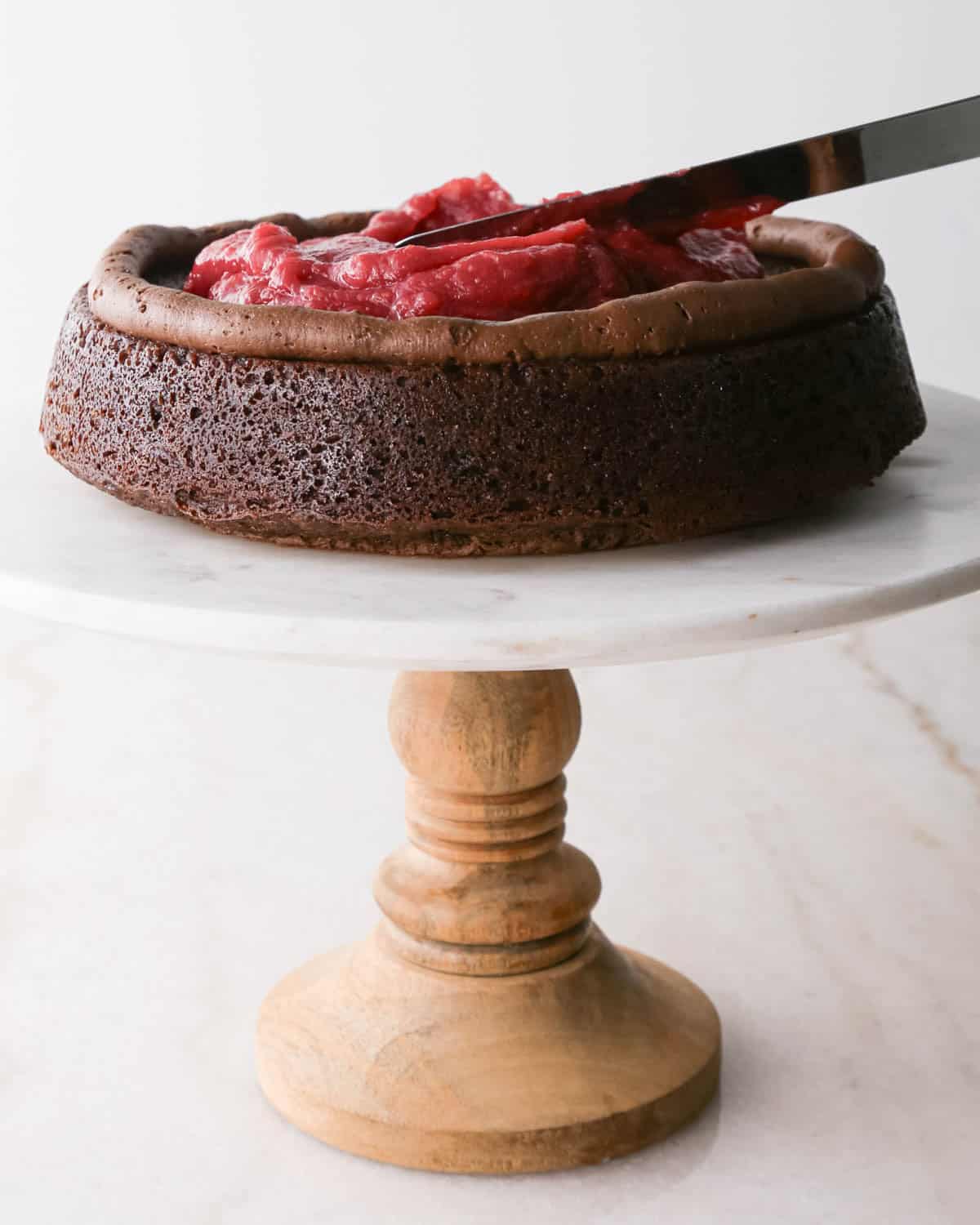 assembling chocolate raspberry cake - raspberry filling being spread into a ring of chocolate frosting on the first layer of cake on a white marble cake stand with a wood base