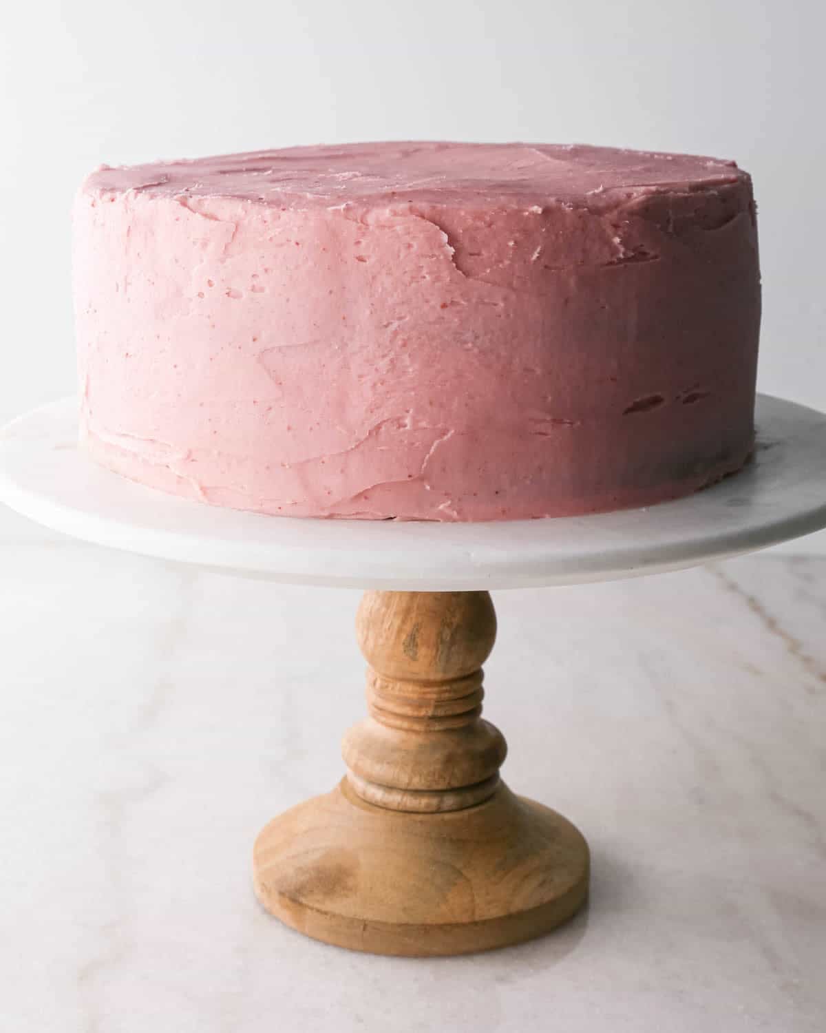 assembling chocolate raspberry cake - raspberry frosting spread all over the cake on a white marble cake stand with a wood base
