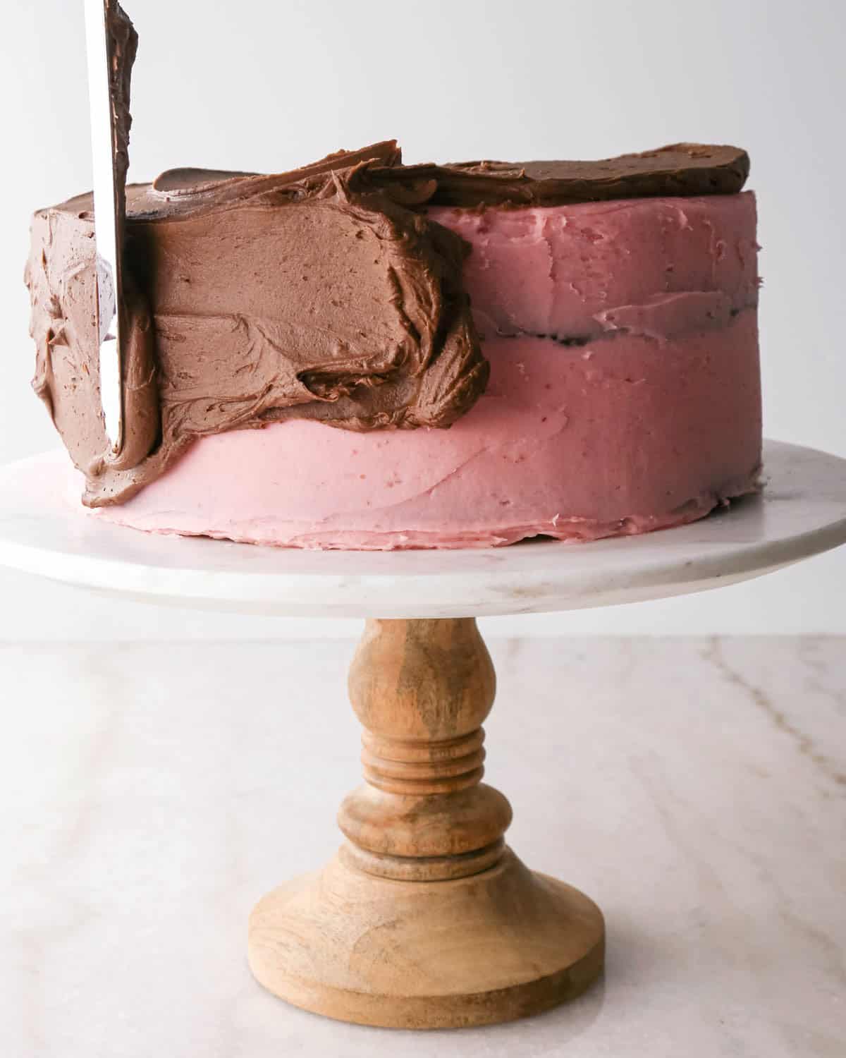 assembling chocolate raspberry cake - chocolate frosting being spread over the raspberry frosting on the cake on a white marble cake stand with a wood base