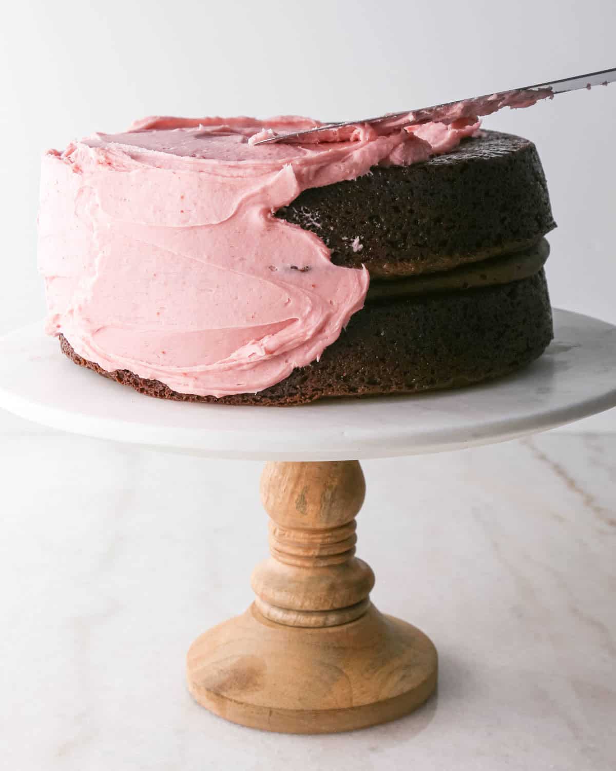 assembling chocolate raspberry cake - raspberry frosting being spread over the cake on a white marble cake stand with a wood base