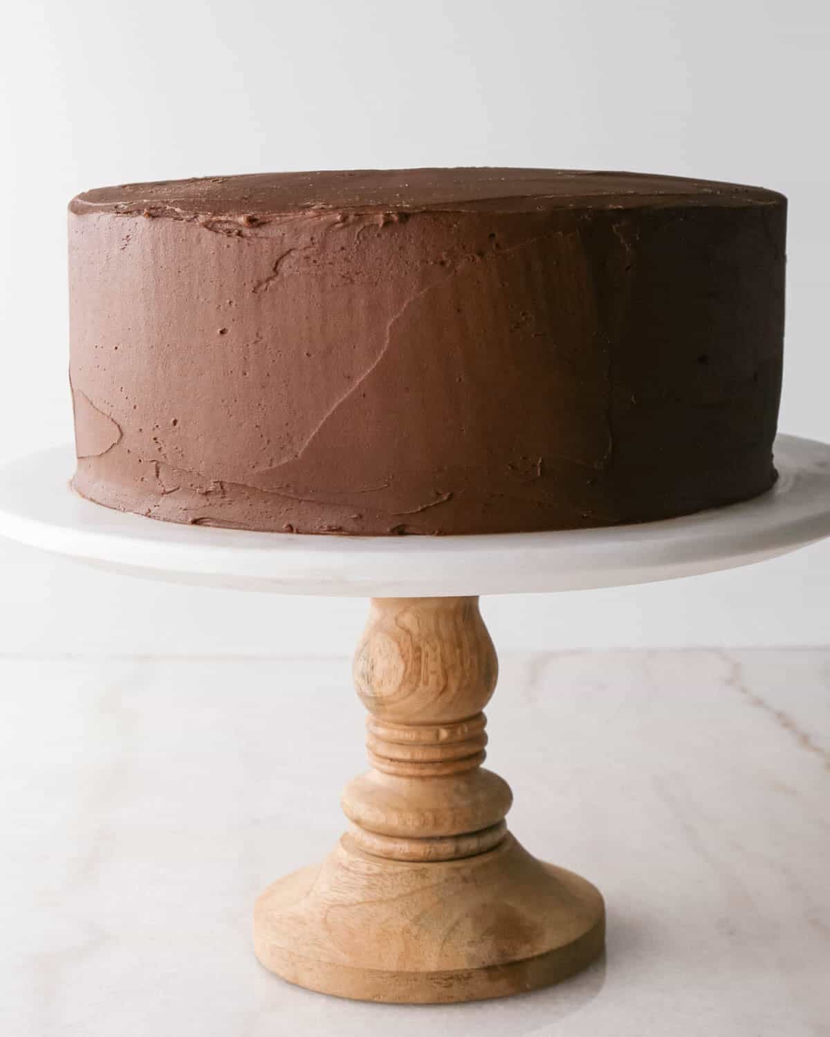 assembling chocolate raspberry cake - chocolate frosting spread over the raspberry frosting on the cake on a white marble cake stand with a wood base