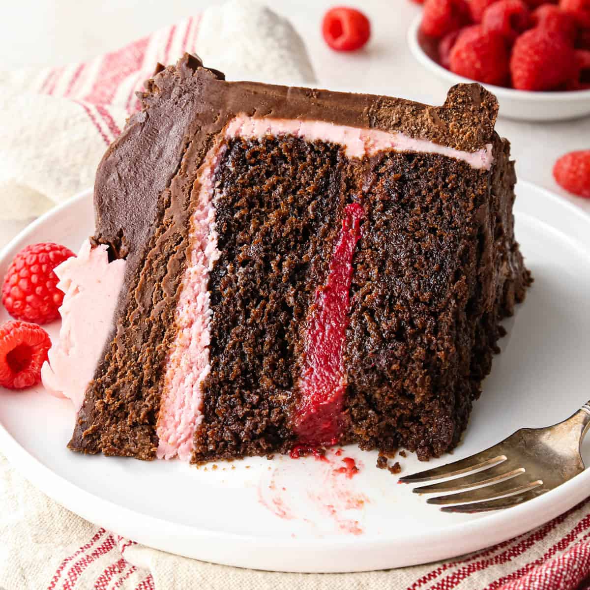 a slice of chocolate raspberry cake on a plate with a bite taken out of it so the moist texture of the cake is visible and there is a dark raspberry layer between the two layers of cake, then a layer of raspberry frosting beneath a thicker layer of chocolate frosting. There's a fork and raspberries on the plate and raspberries and a red and white striped towel int he background