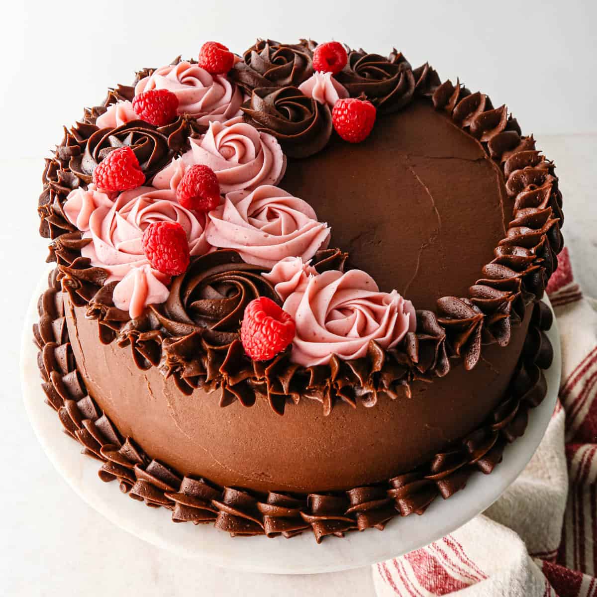 chocolate raspberry cake on a white cake plate decorated with both chocolate roses and raspberry frosting roses, and fresh raspberries