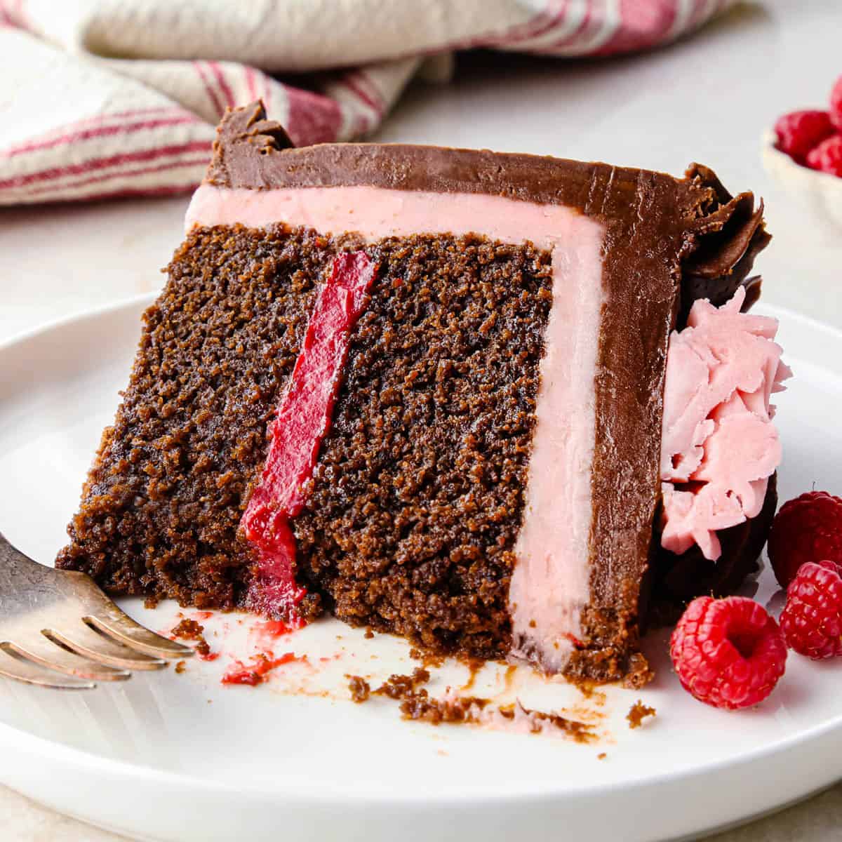 a slice of chocolate raspberry cake on a plate with ab ite taken out so the moist texture of the cake is visible and there is a dark raspberry layer between the two layers of cake, then a layer of raspberry frosting beneath a thicker layer of chocolate frosting. There's a fork and raspberries on the plate and raspberries and a red and white striped towel int he background