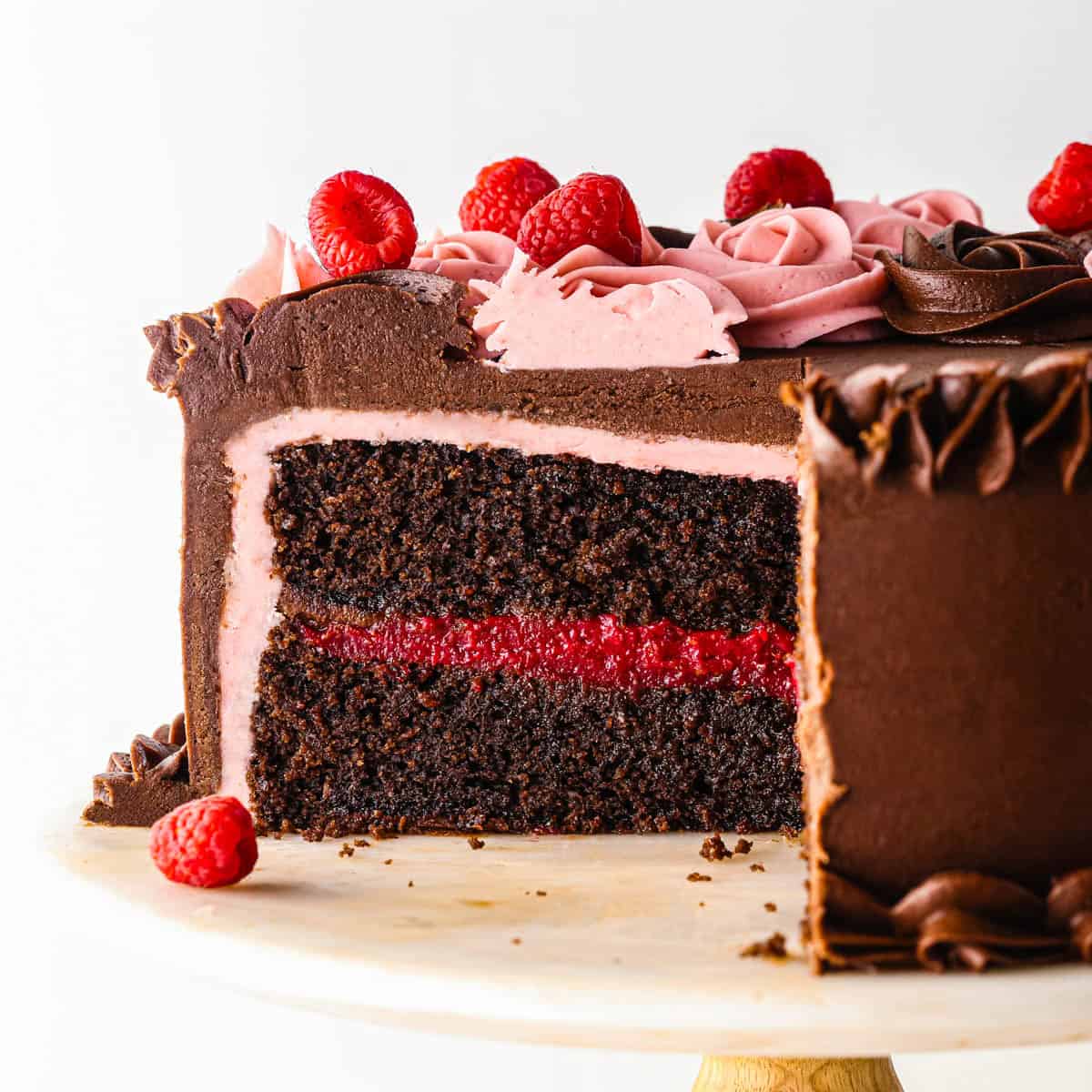 chocolate raspberry cake on a white cake plate, a piece has been cut out so you can see the two layers moist chocolate cake and dark raspberry filling in between. You can also see the bottom layer of raspberry frosting, top and of chocolate buttercream adn the frosting roses and fresh raspberries on top