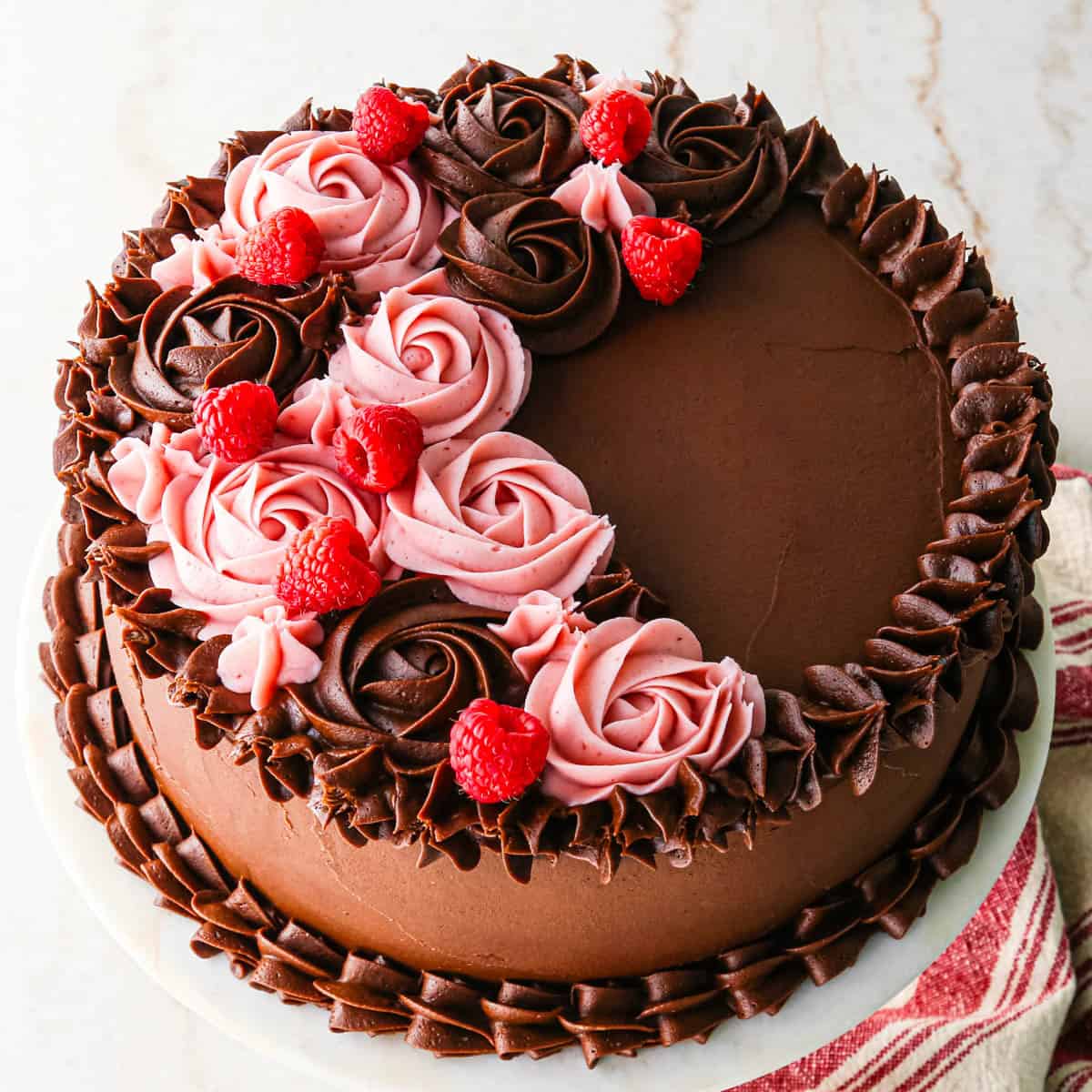 chocolate raspberry cake on a white cake plate decorated with both chocolate roses and raspberry frosting roses, and fresh raspberries