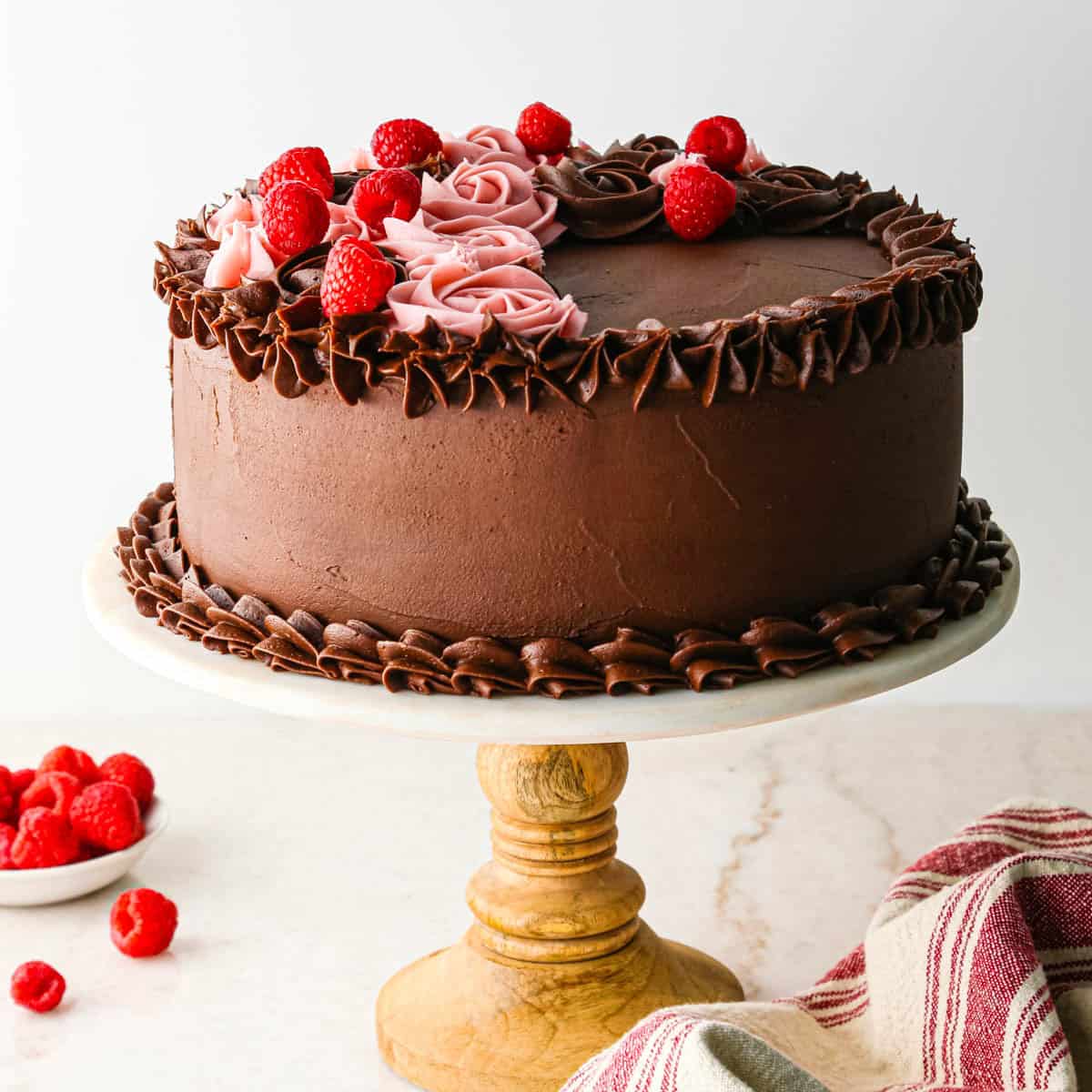 chocolate raspberry cake on a white and wood cake stand decorated with both chocolate roses and raspberry frosting roses, and fresh raspberries