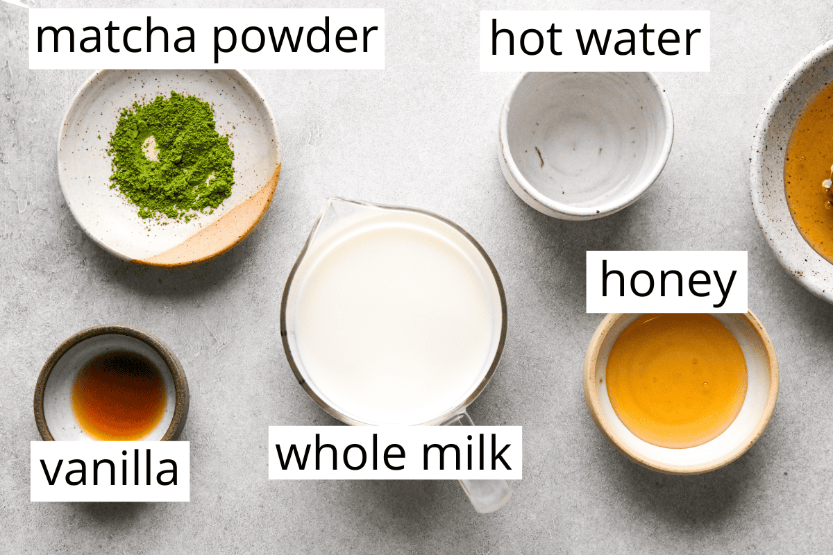 overhead photo of the ingredients in this matcha latte recipe in bowls and labeled matcha powder, hot water, honey, whole milk and vanilla