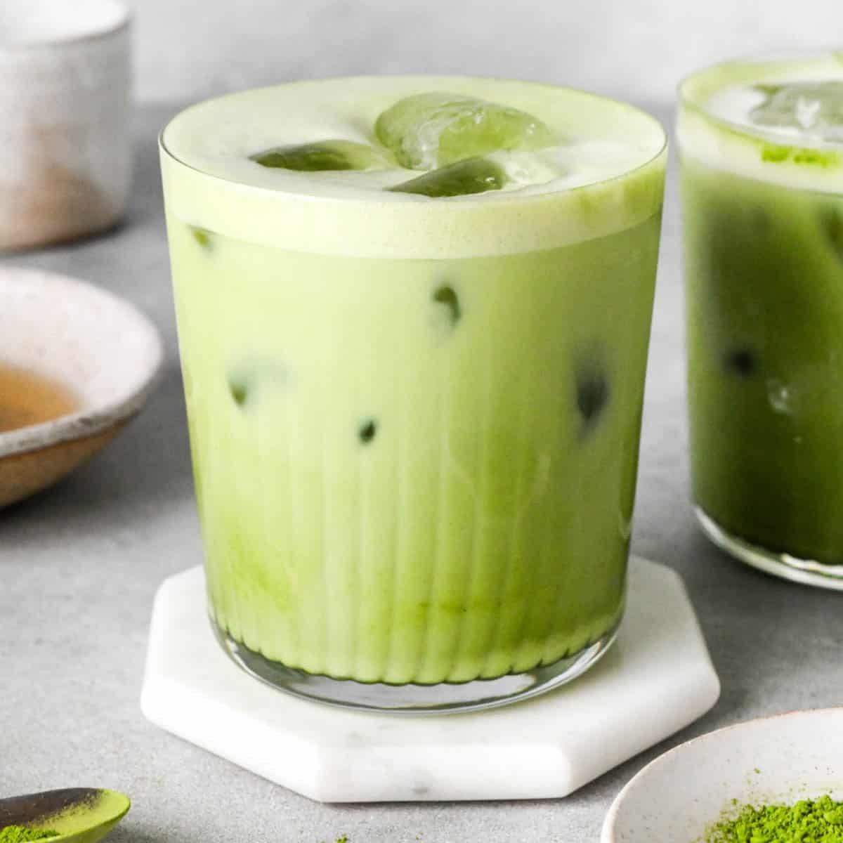 front view of an iced matcha latte in a glass mug with a layer of foamy at the top and another matcha iced latte to the right of it