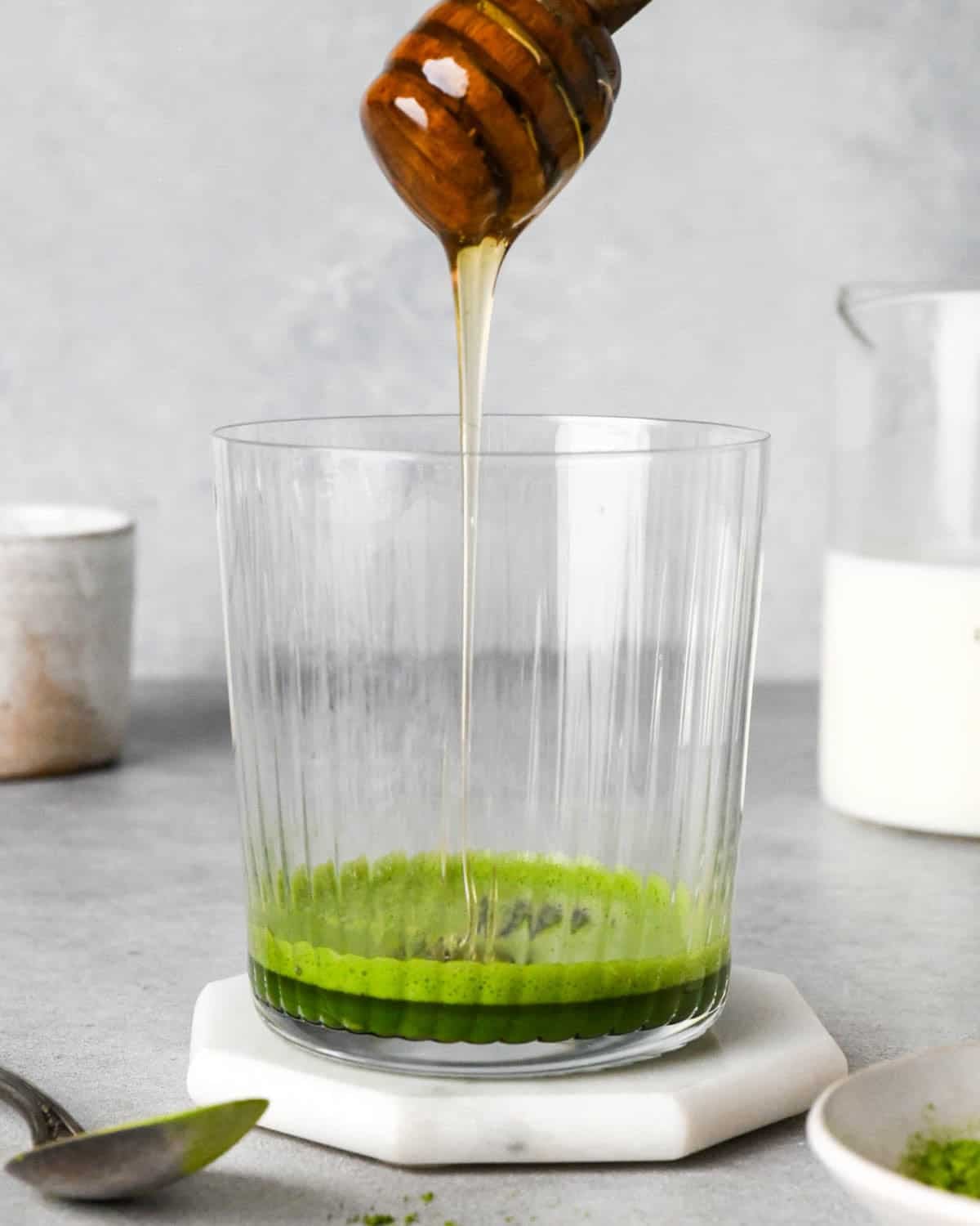 photo showing how to make an iced matcha latte - front view of honey being poured into a glass cup with matcha and water at the bottom