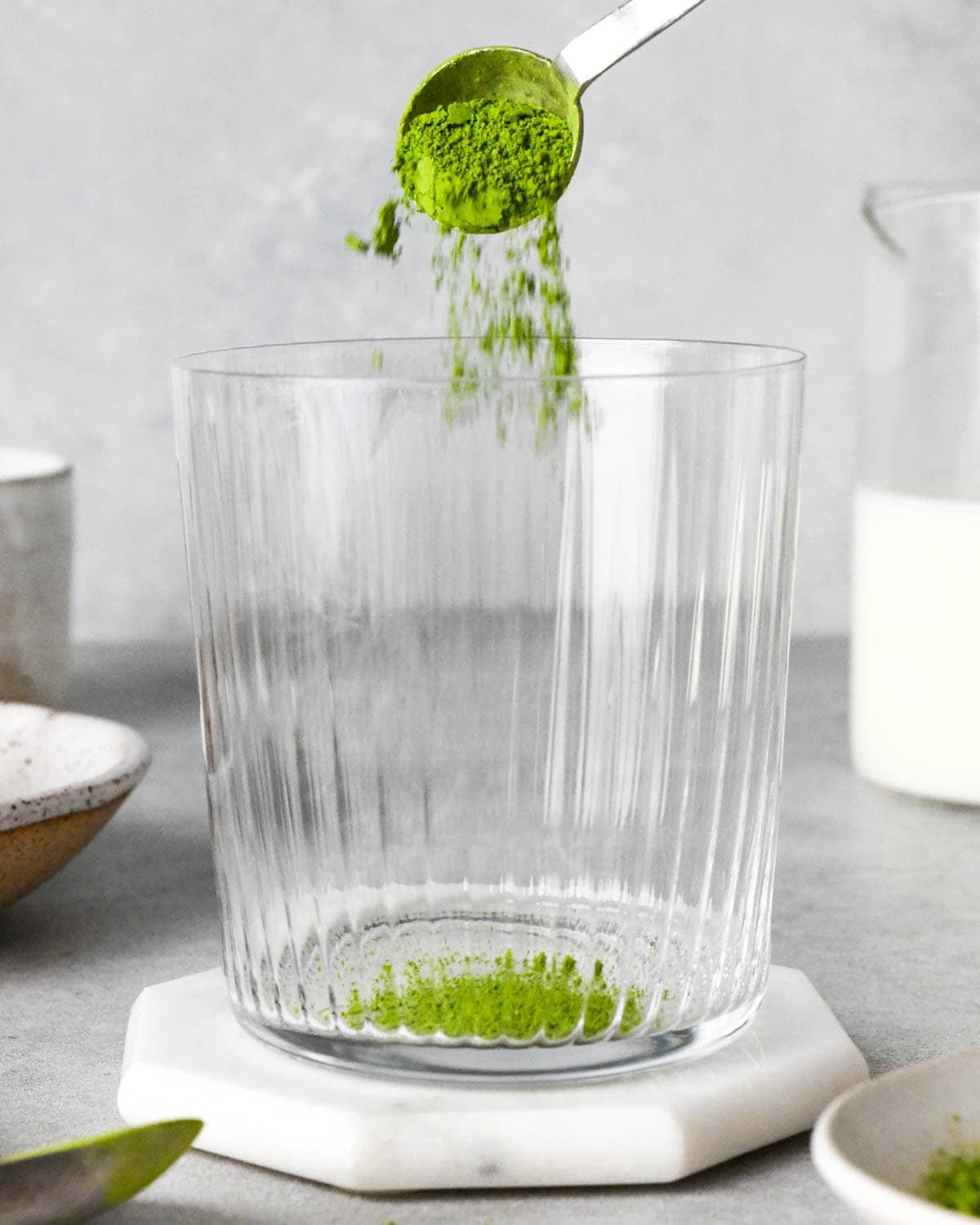 making an iced matcha latte - matcha powder being poured into a glass cup from a silver measuring spoon