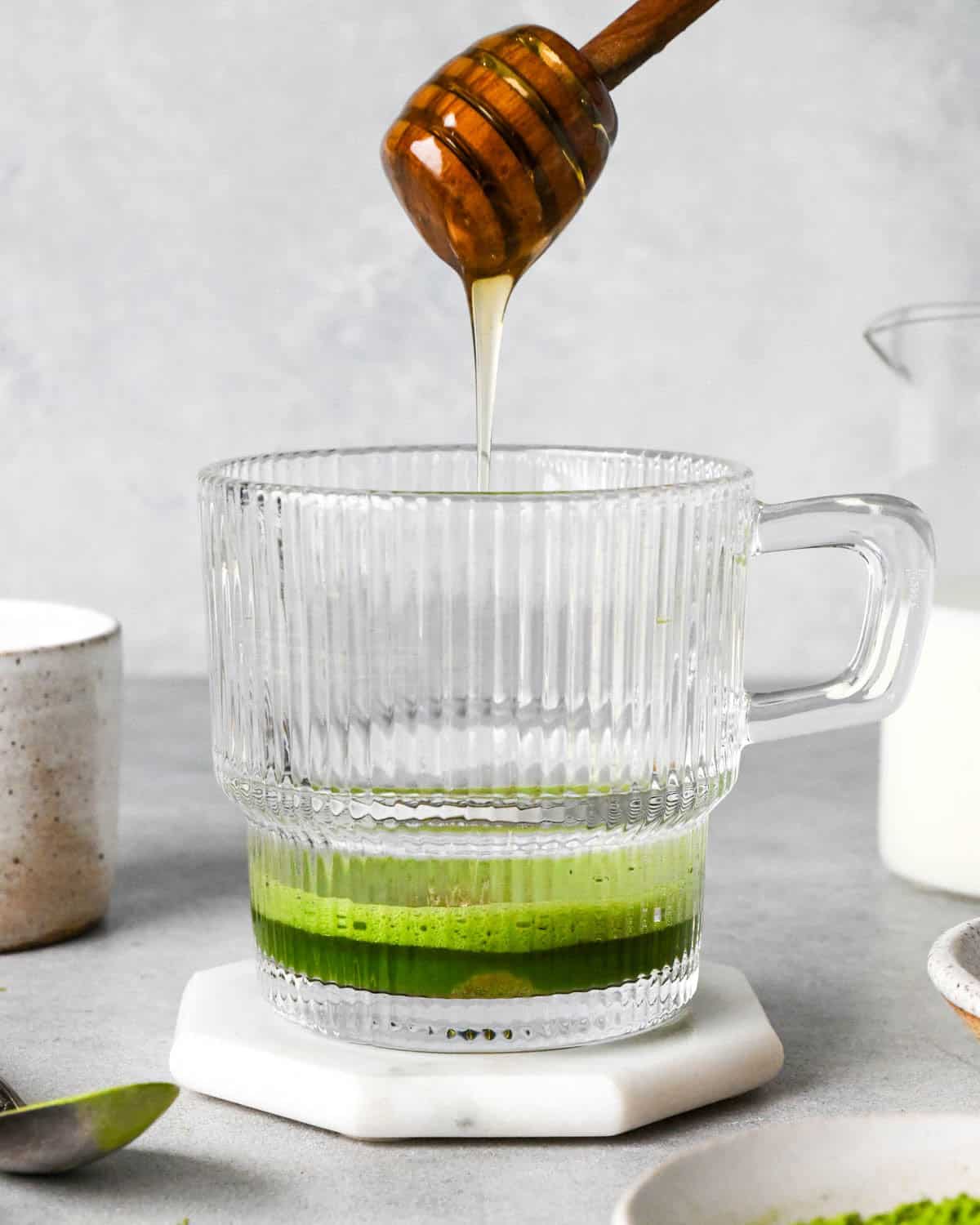 photo showing how to make a matcha latte - front view of honey being poured into a glass mug with matcha powder and hot water