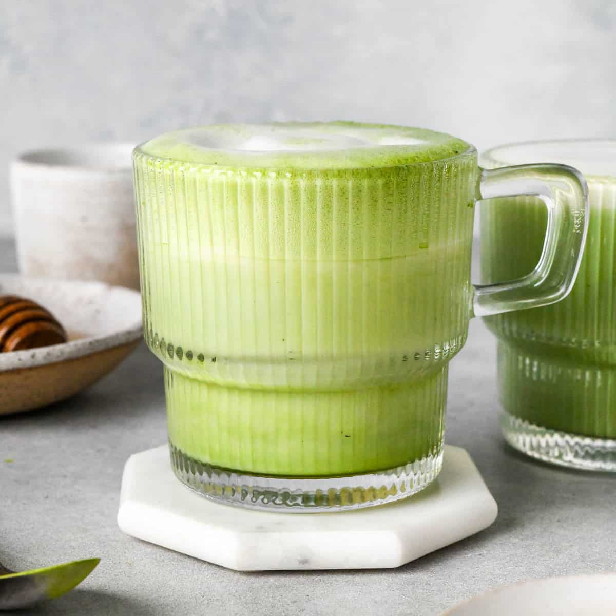 matcha latte in a clear glass mug with a handle with foamy milk on top and another matcha latte to the back and right of it and a bowl of honey to the left