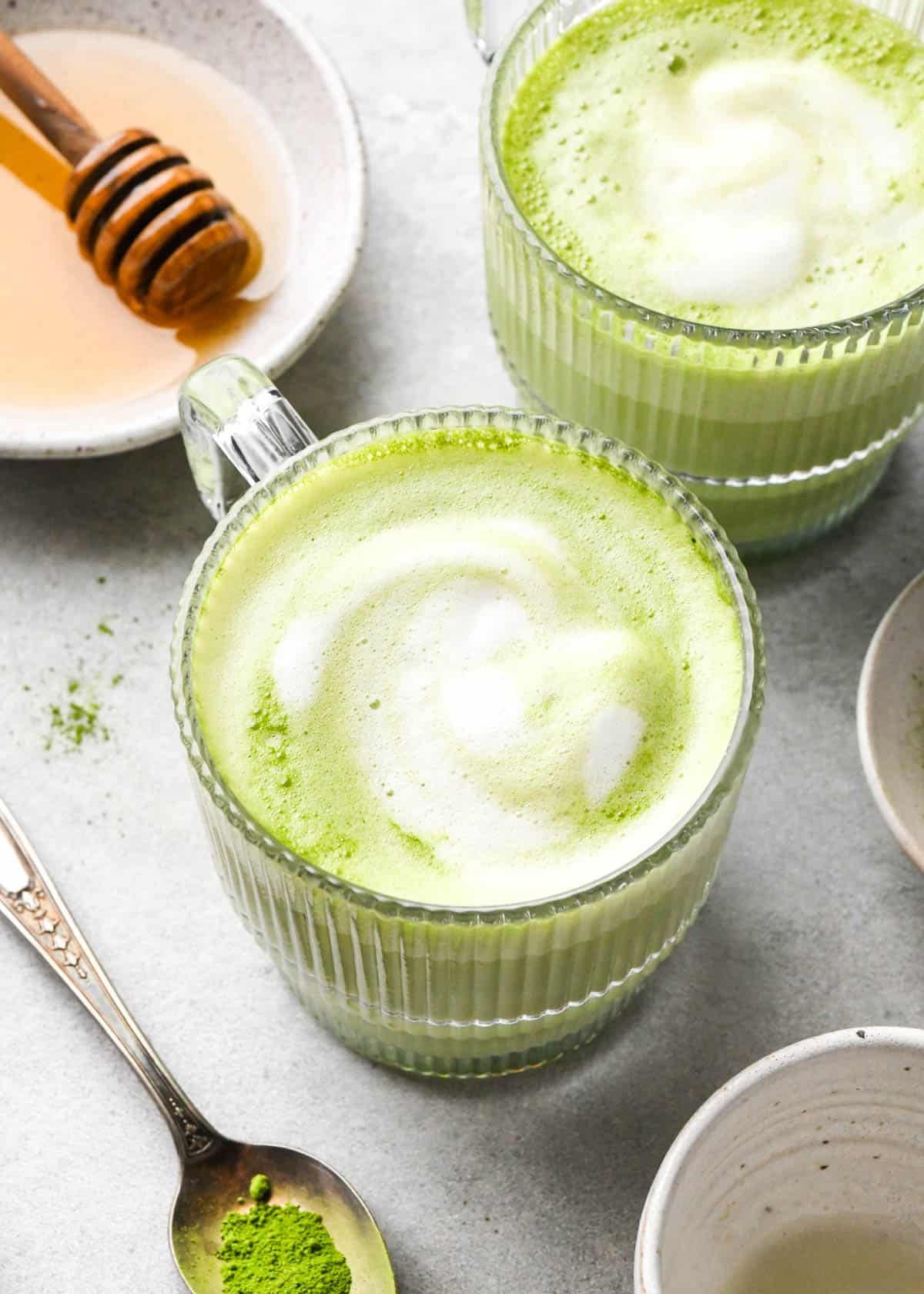 matcha latte in a clear glass mug with a handle with foamy milk on top and another matcha latte to the back and right of it